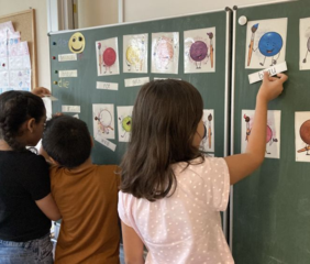 Drei Kinder betrachten im Klassenzimmer eine grüne Tafel mit Zeichnungen; ein Mädchen greift nach oben
