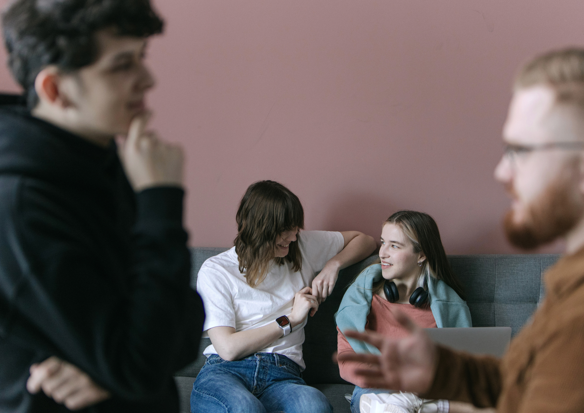 Vier junge Menschen im Gespräch: zwei lachen auf einem grauen Sofa, zwei stehen unscharf im Vordergrund