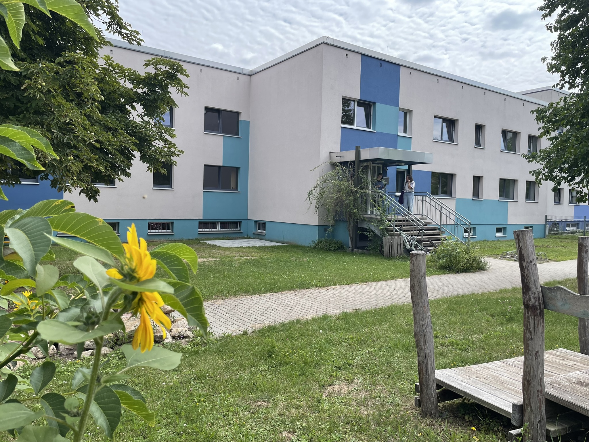 Blau-weiß gestrichenes Gebäude mit Eingangstreppe und zwei Personen davor, im Vordergrund eine Sonnenblume