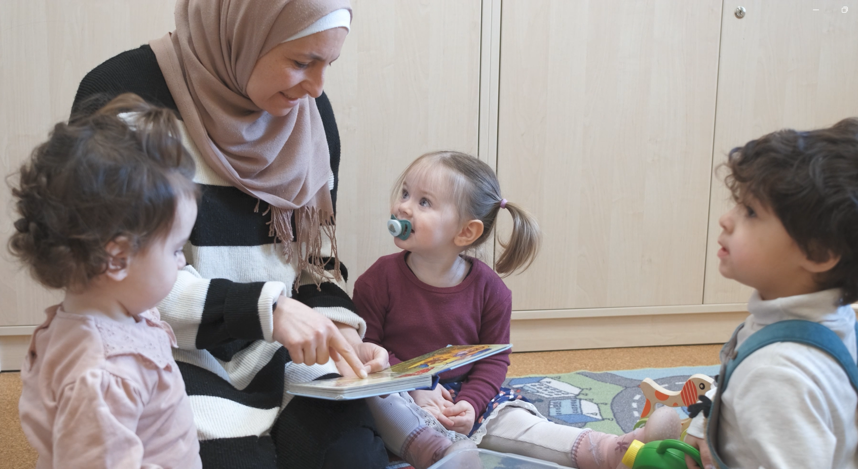 Eine Erzieherin mit Kopftuch liest zwei Kindern aus einem Buch vor.