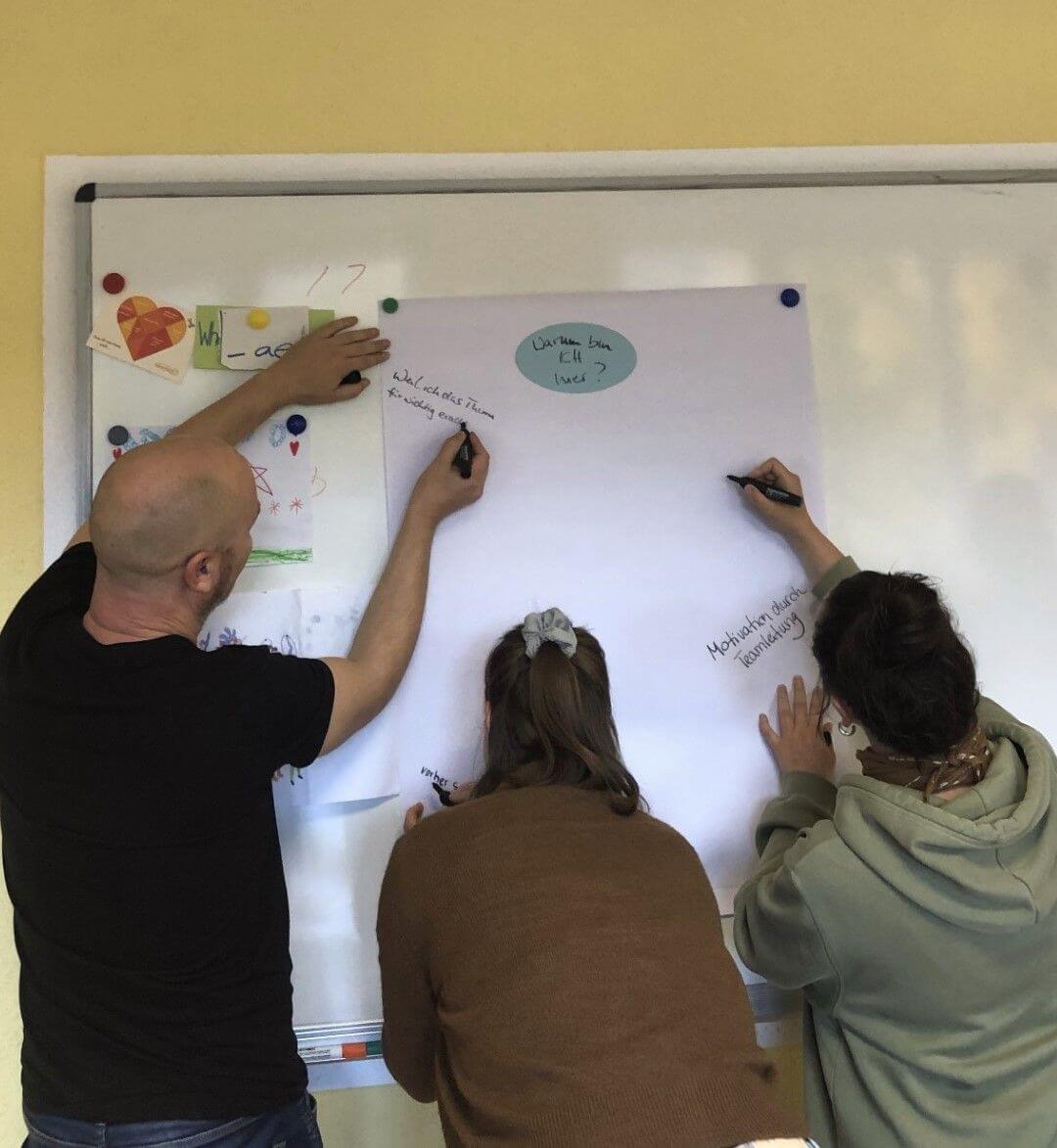 Drei Personen beschriften ein großes Plakat am Whiteboard mit Notizen und Fragen