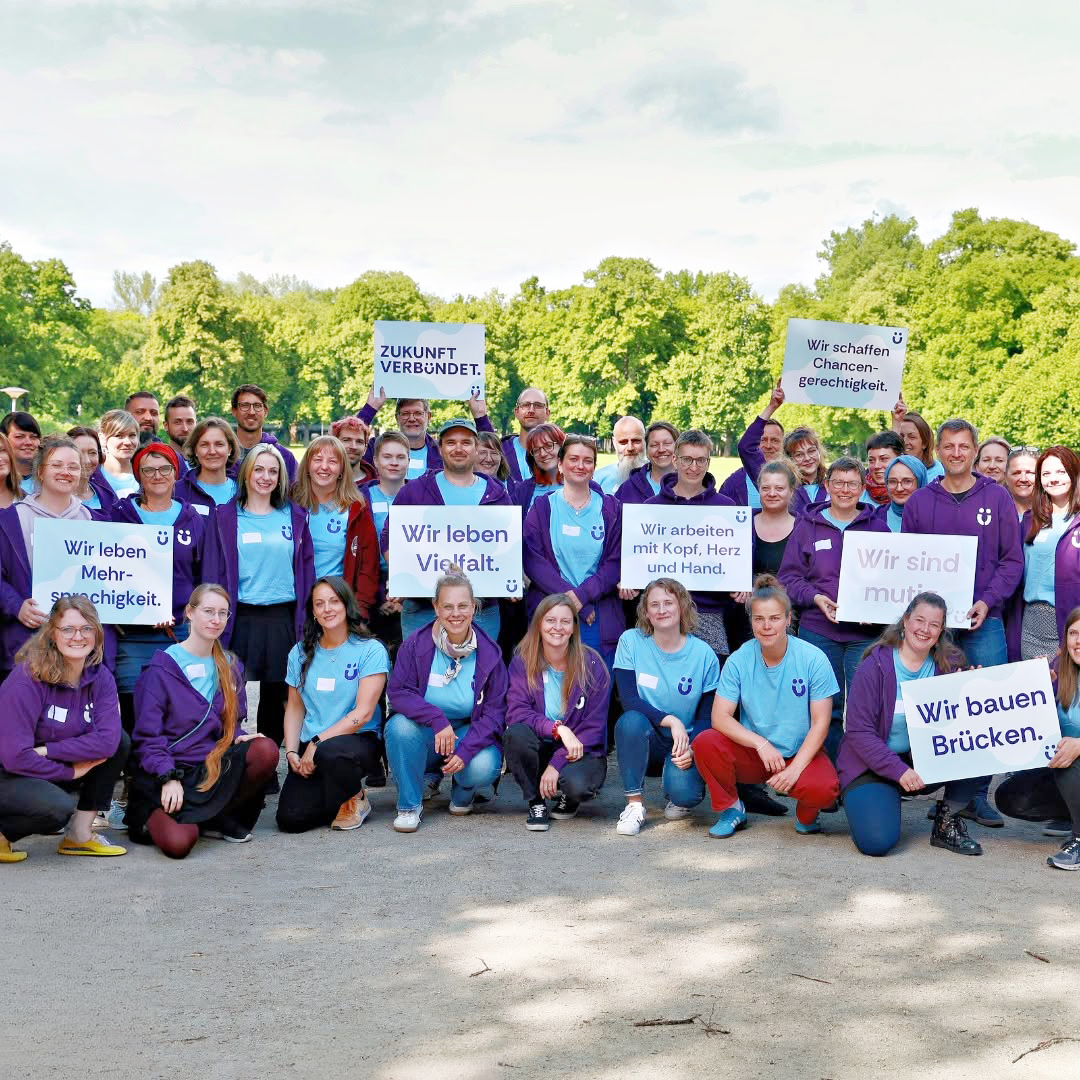 Gruppe von Mitarbeitenden in lila und hellblauen Shirts mit Schildern wie „Wir leben Vielfalt" und „Wir bauen Brücken" im Park