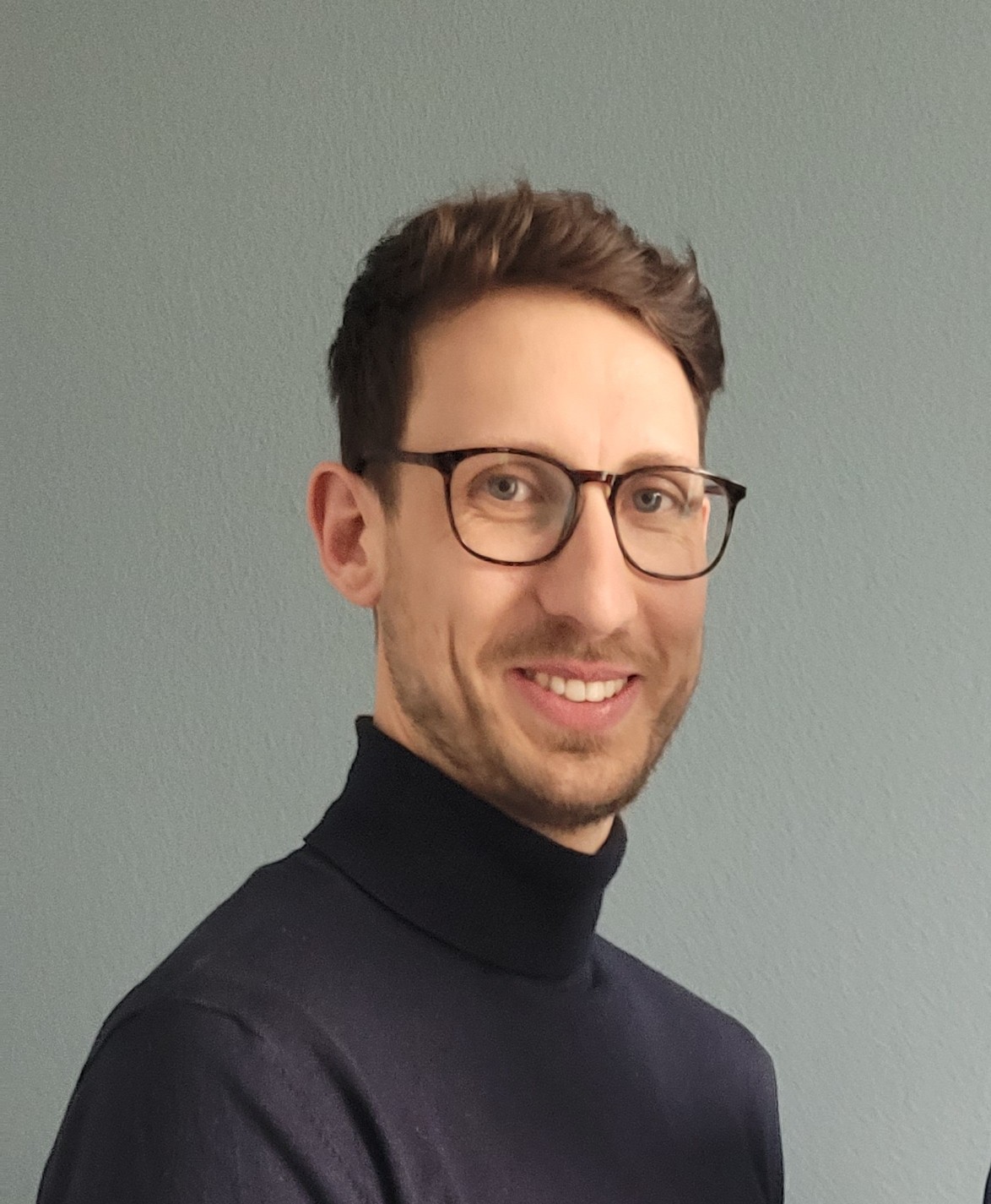 Lächelnder Mann mit Brille und schwarzem Rollkragenpullover vor graugrünem Hintergrund
