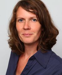 Frau mit braunen, schulterlangen Haaren in dunkelblauem Hemd vor hellem Hintergrund