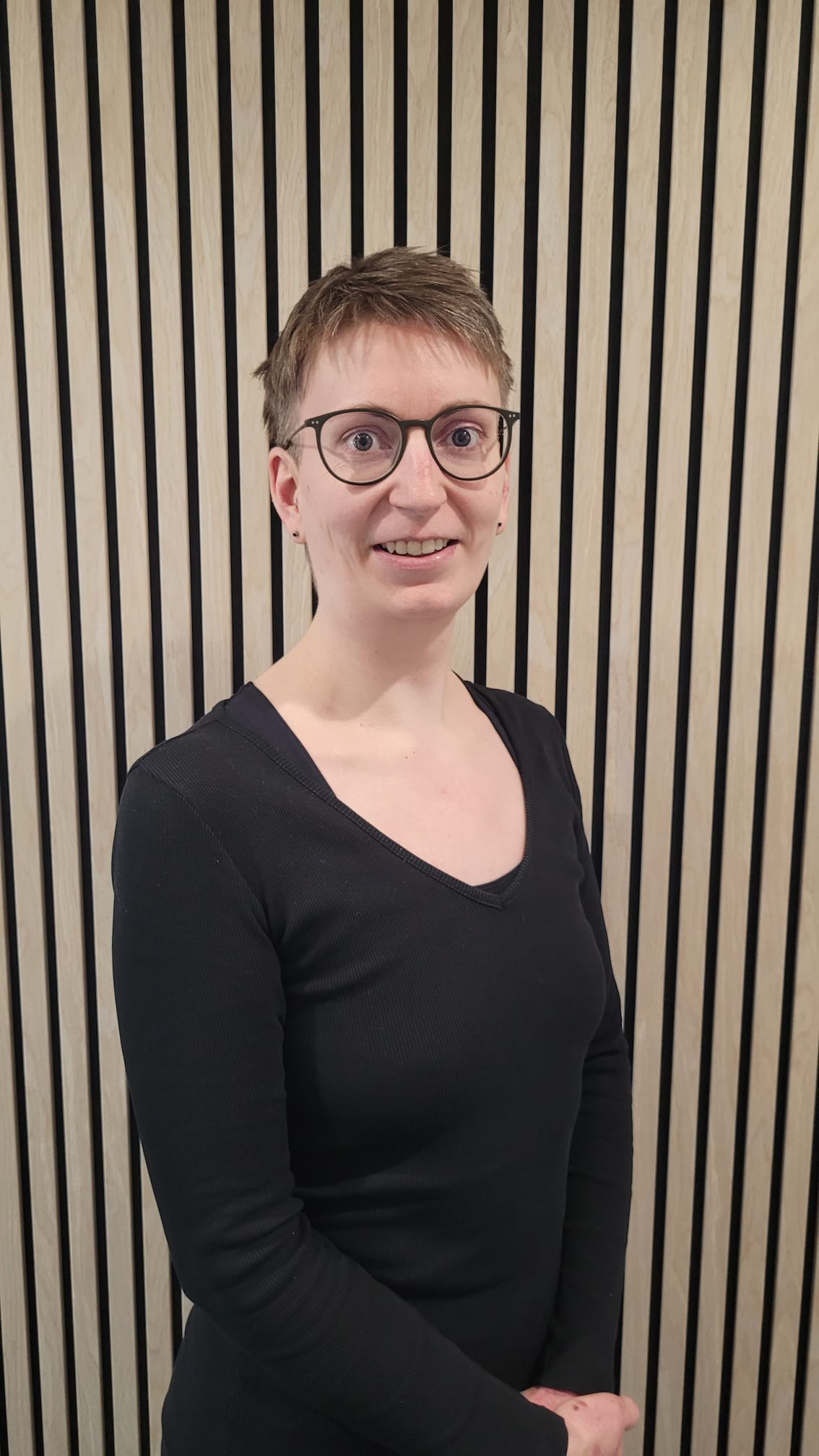 Lächelnde Person mit kurzen blonden Haaren und Brille in schwarzem Langarmshirt vor einer Holzlattenwand
