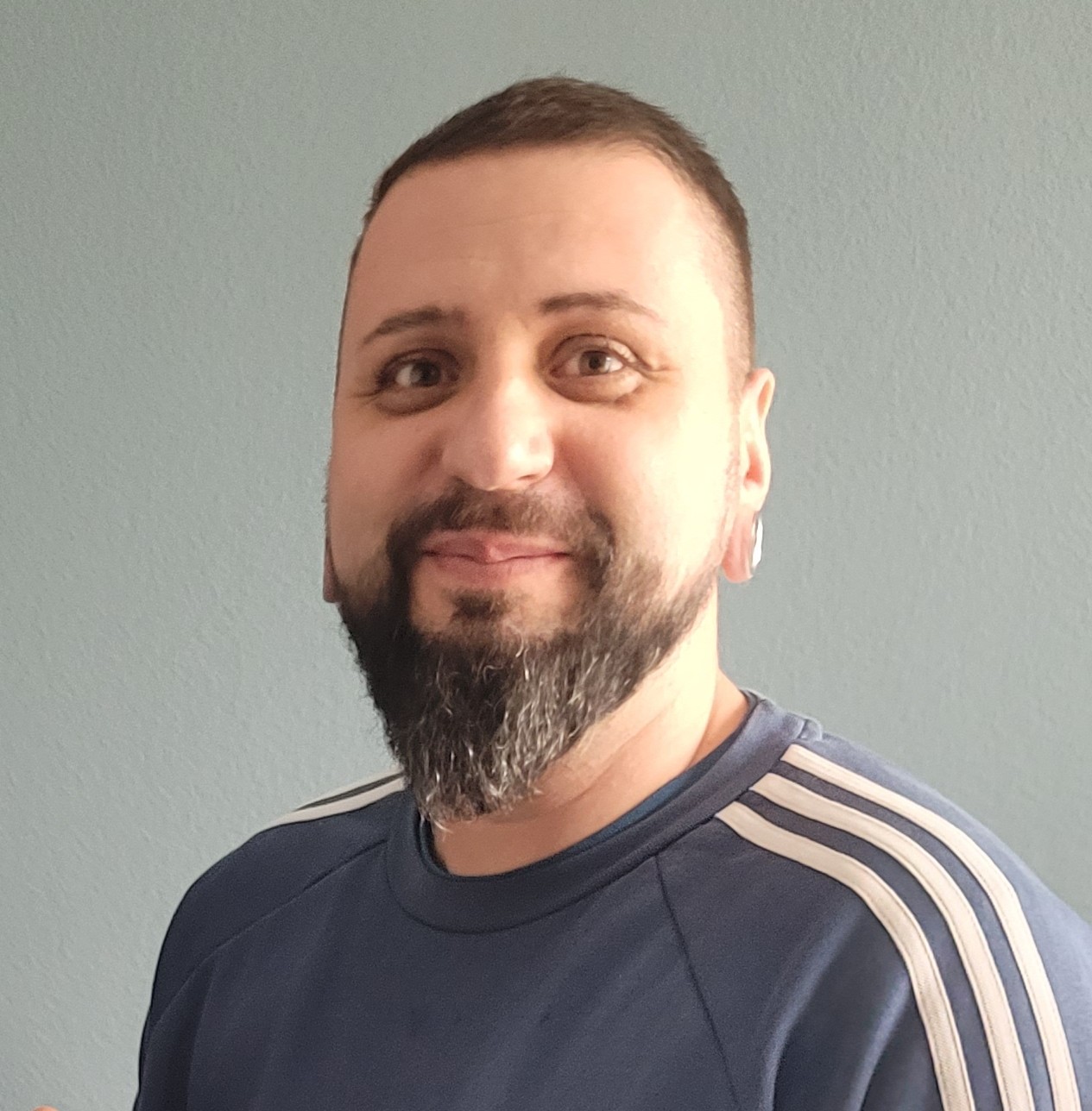 Lächelnder Mann mit kurzem Haar und Vollbart, Ohrring und blauem Shirt mit weißen Streifen