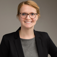 Lächelnde Frau mit Brille, schwarzem Blazer und grauem Shirt vor grauem Hintergrund