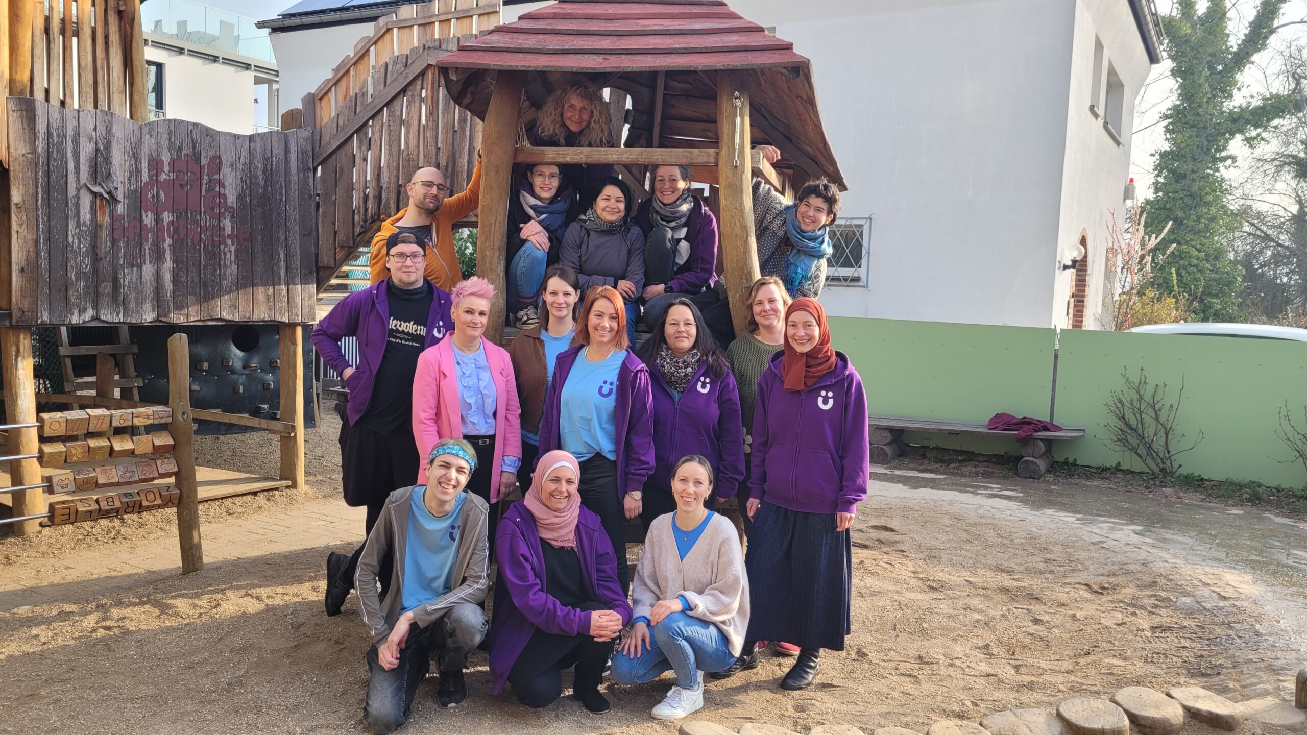 Gruppenfoto unseres Kita‑Teams auf dem Spielplatz vor der Holzspielanlage.