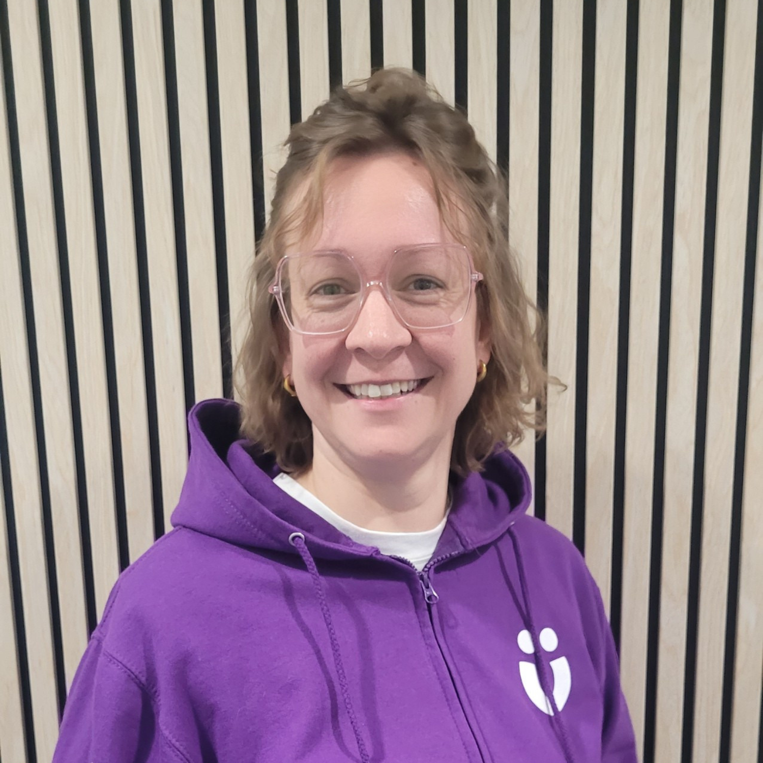 Lächelnde Person mit Brille und lockigem Haar in lila Kapuzenpullover vor einer Holzlattenwand