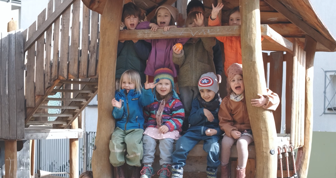 Kinder sitzen und stehen winkend auf einem hölzernen Spielgerüst