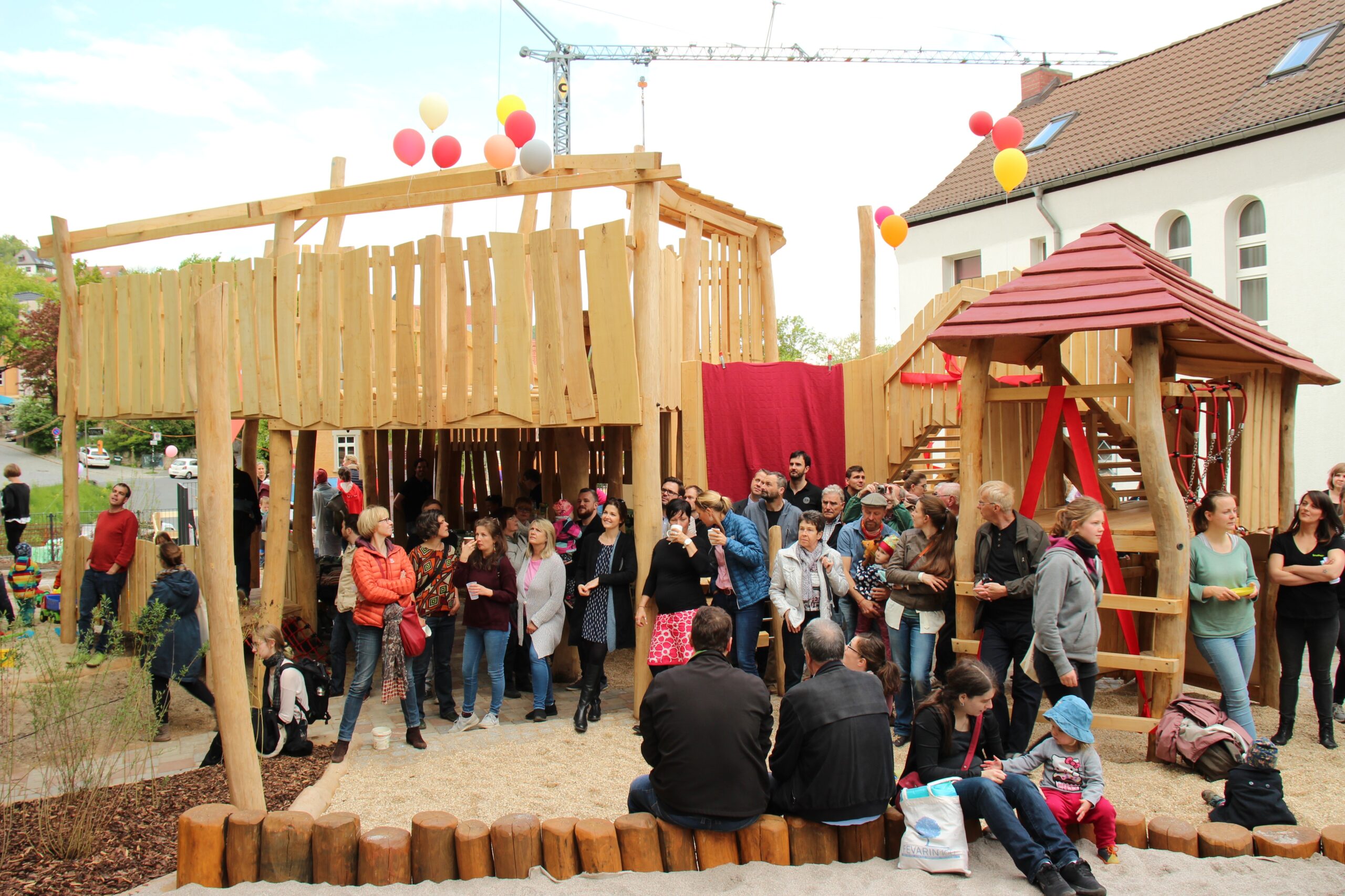 Zahlreiche Erwachsene und Kinder versammeln sich an einem neuen Holzspielplatz mit bunten Luftballons