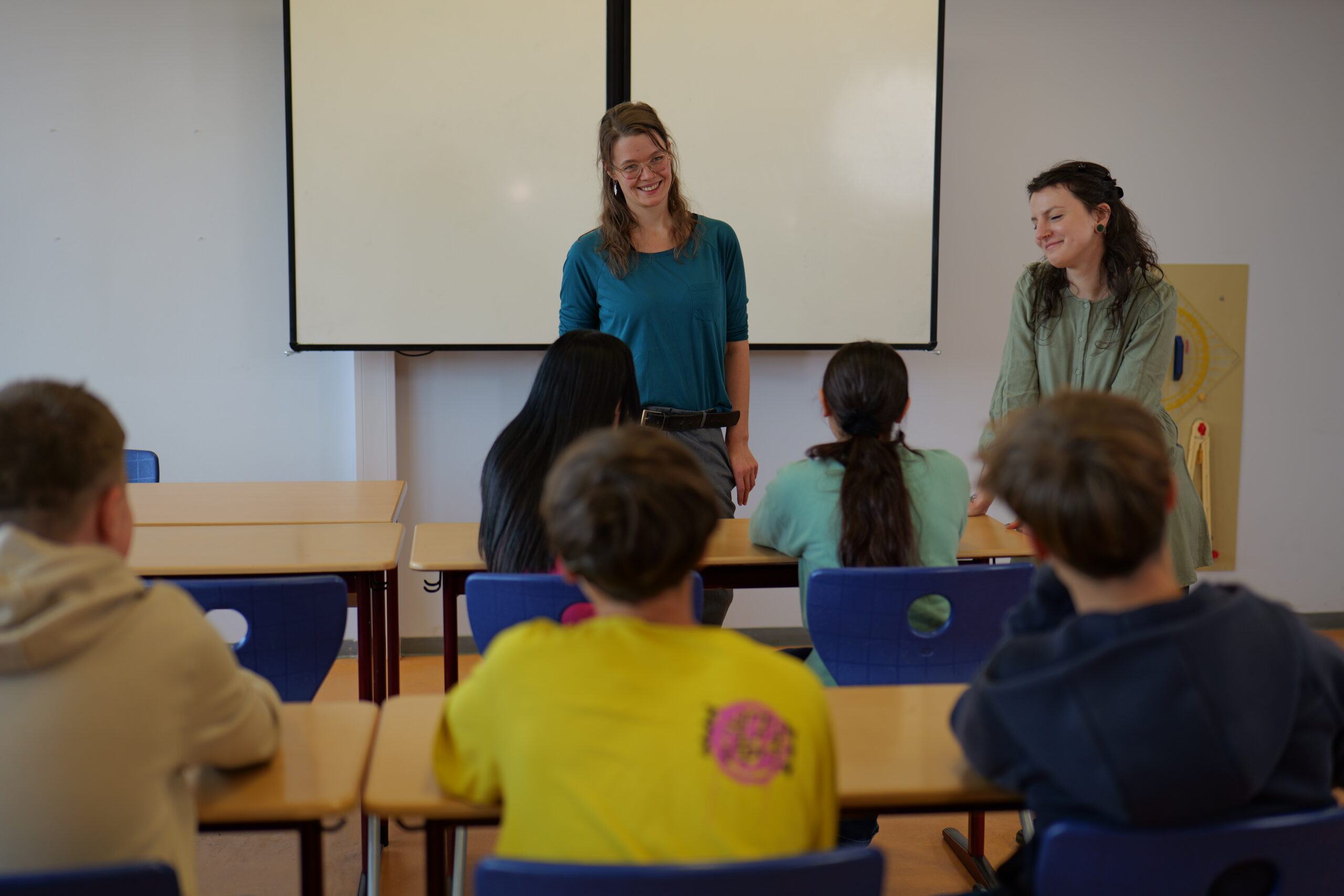 Zwei Frauen stehen lächelnd vor einer Schulklasse, die an Tischen im Klassenzimmer sitzt