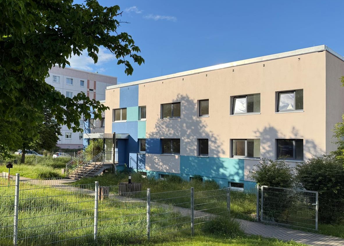 Zweigeschossiges Gebäude mit beiger Fassade und blauen Akzenten, davor Zaun und Wiese, links ein Wohnblock