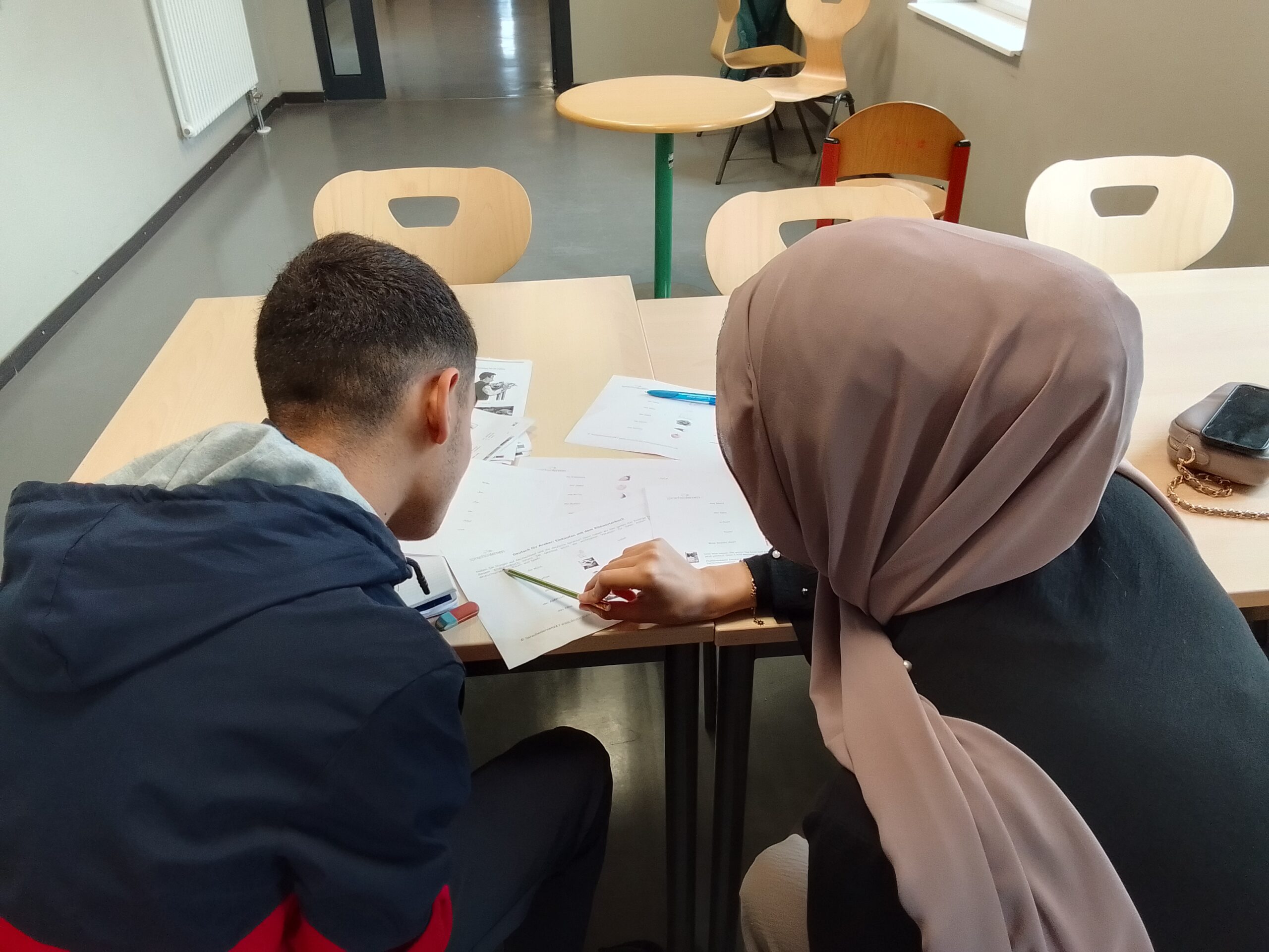 Ein junger Mann und eine Frau mit Kopftuch sitzen an einem Tisch und bearbeiten gemeinsam Arbeitsblätter