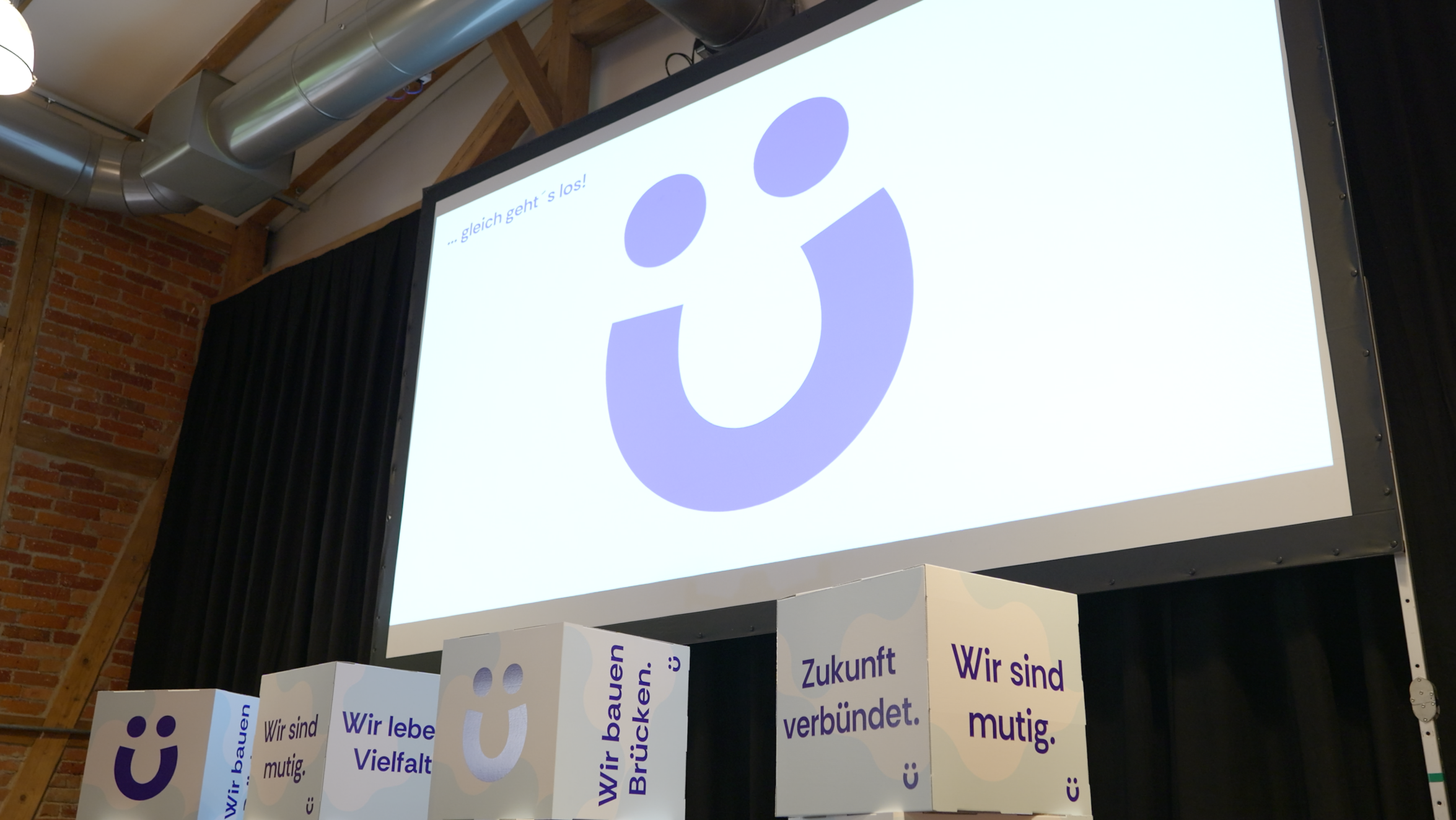 Leinwand mit lila Smiley-Logo und Schriftzug … gleich geht's los!, davor Sitzwürfel mit Slogans wie "Zukunft verbündet", "Wir sind mutig" und "Wir bauen Brücken"