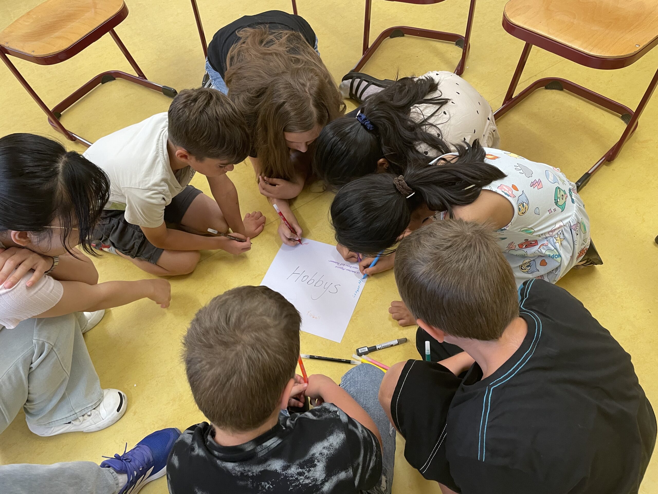 Kinder sitzen im Kreis auf dem Boden und schreiben gemeinsam auf ein Blatt Papier mit der Überschrift Hobbys