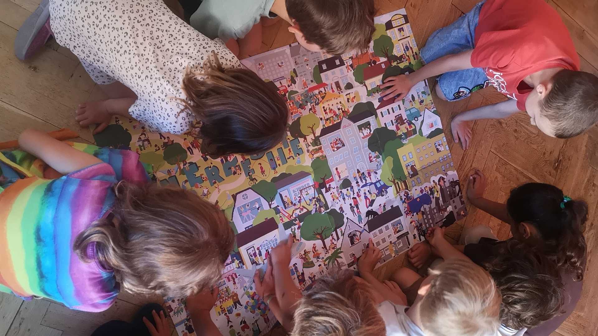 Mehrere Kinder knien und liegen auf dem Holzfußboden um eine große illustrierte Spielmatte mit einer Stadt