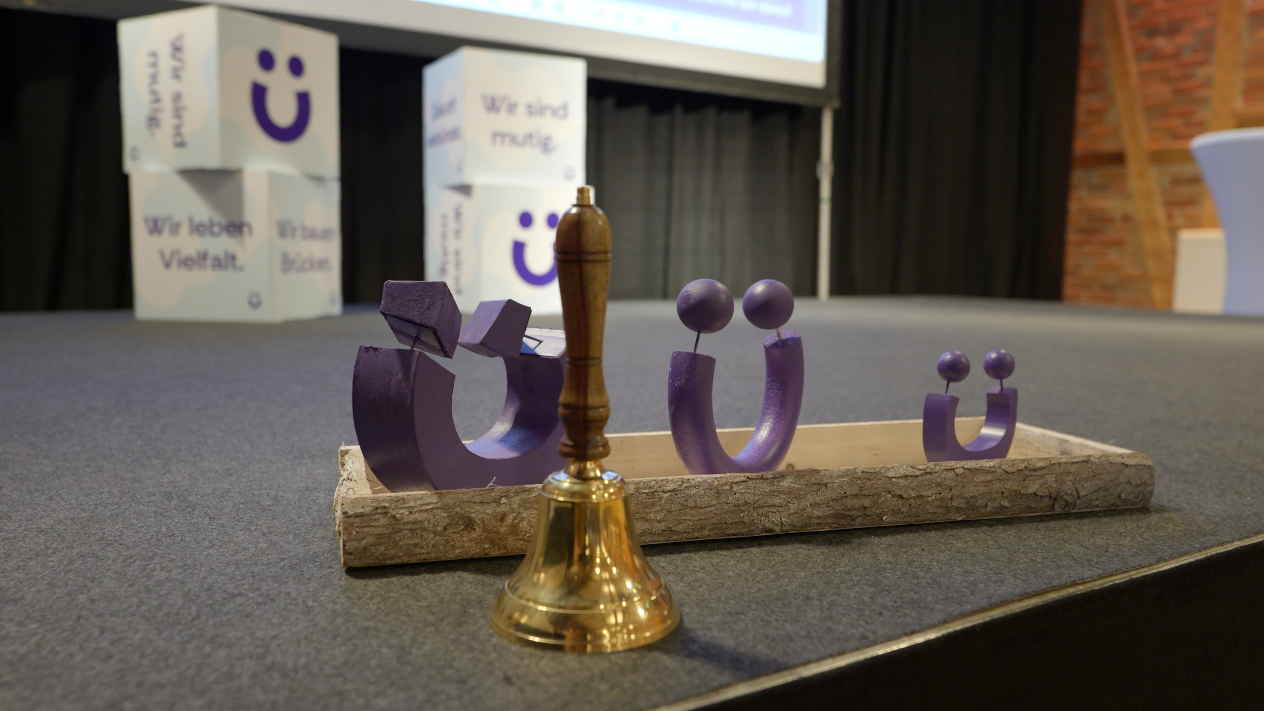 Goldene Tischglocke und drei violette, unterschiedlich große Figuren auf einem Holztablett; im Hintergrund Bühne mit Würfeln und Aufschriften wie Wir leben Vielfalt