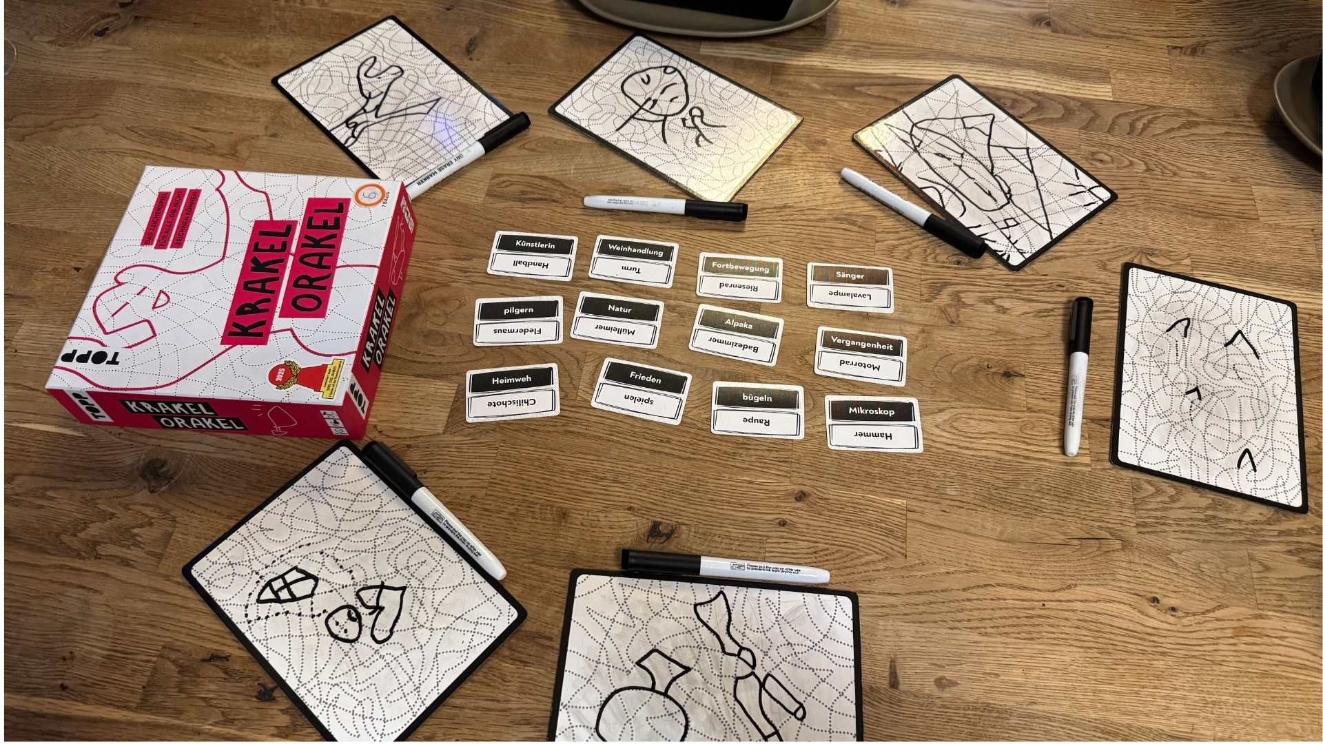 Spiel Krakel Orakel auf einem Holztisch: Schachtel, Begriffskarten, Stifte und Tafeln mit Kritzelzeichnungen