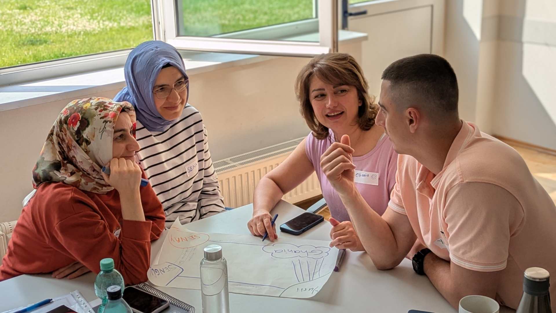 Vier Personen sitzen an einem Tisch und diskutieren bei einer Gruppenarbeit mit Notizen auf einem Plakat