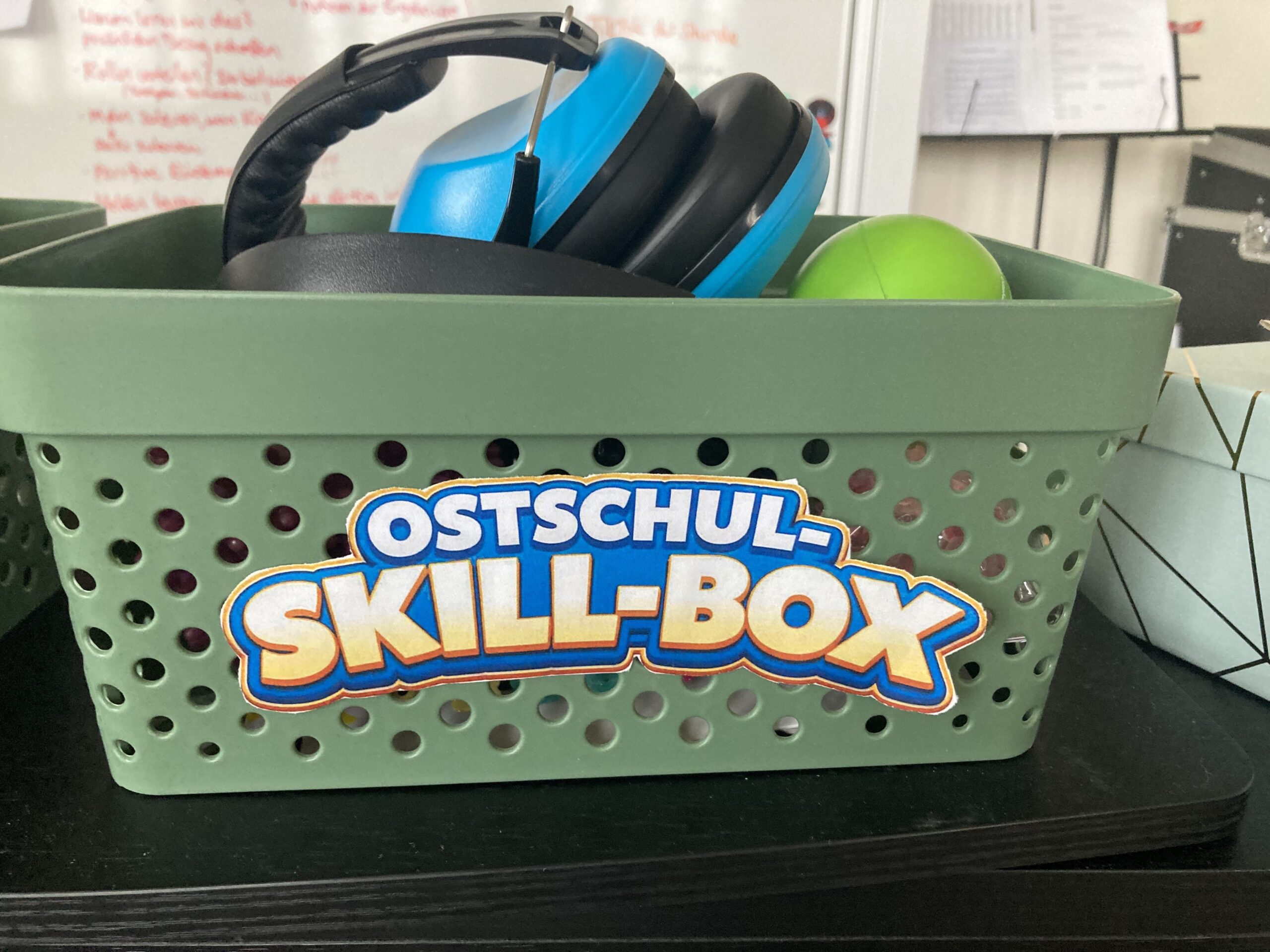 Grüner Korb mit der Aufschrift Ostschul-Skill-Box, darin ein Kopfhörer und ein grüner Ball