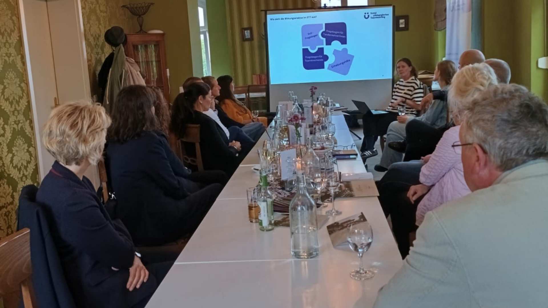 Personen sitzen an einer langen Tafel, während eine Präsentation zur Wirkungsweise im ETT gezeigt wird