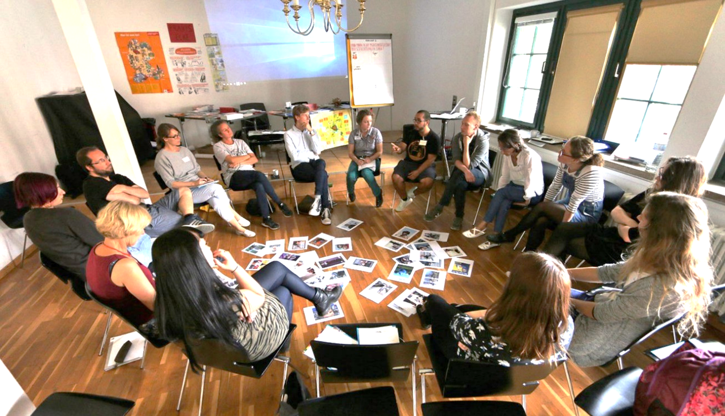 Gruppe von Menschen sitzt in einem Stuhlkreis, in der Mitte liegen Fotos auf dem Boden