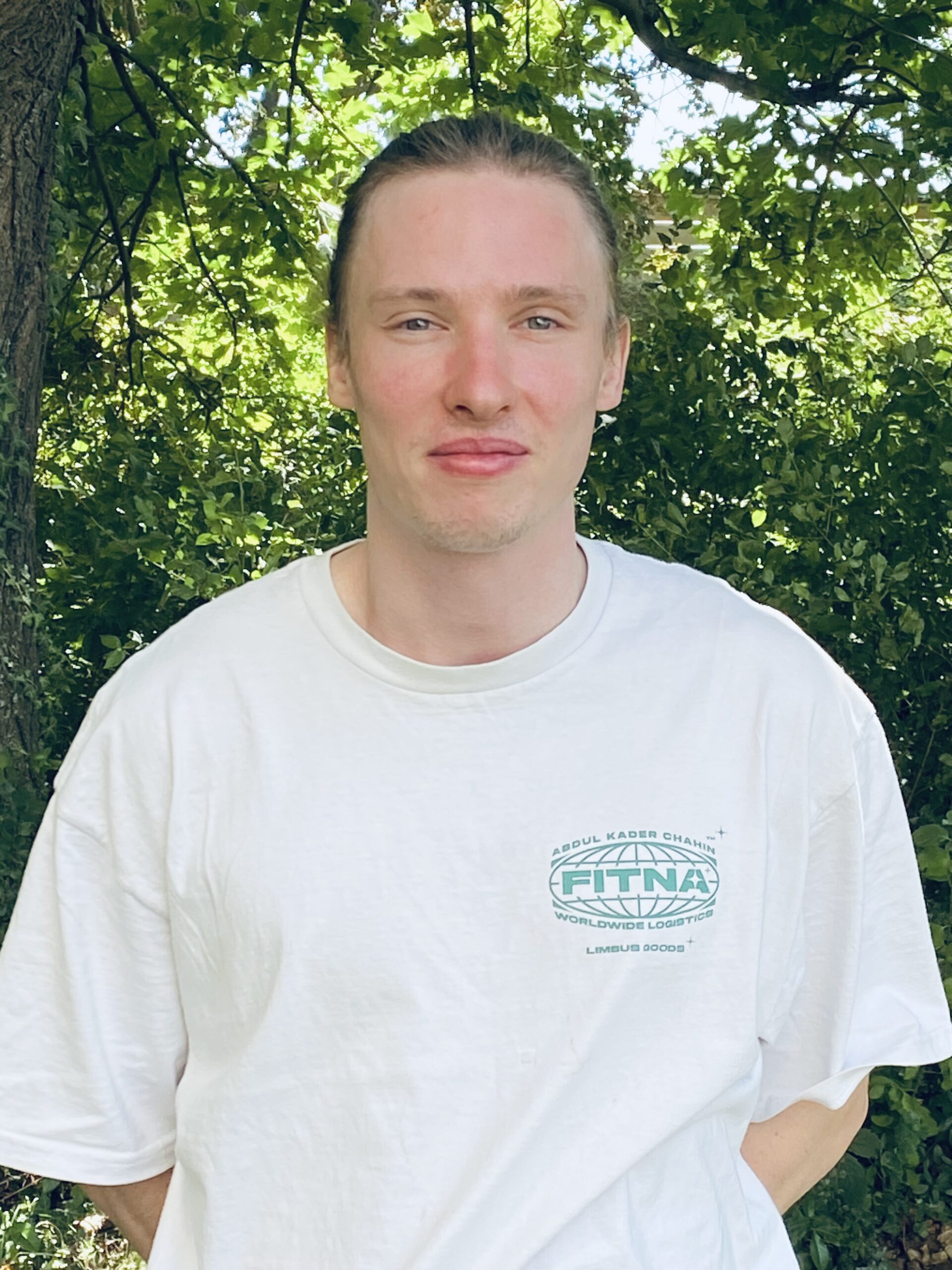 Ein Mann mit zurückgekämmten Haaren trägt ein weißes T-Shirt mit Firmenlogo und steht lächelnd im Freien vor grünem Laub