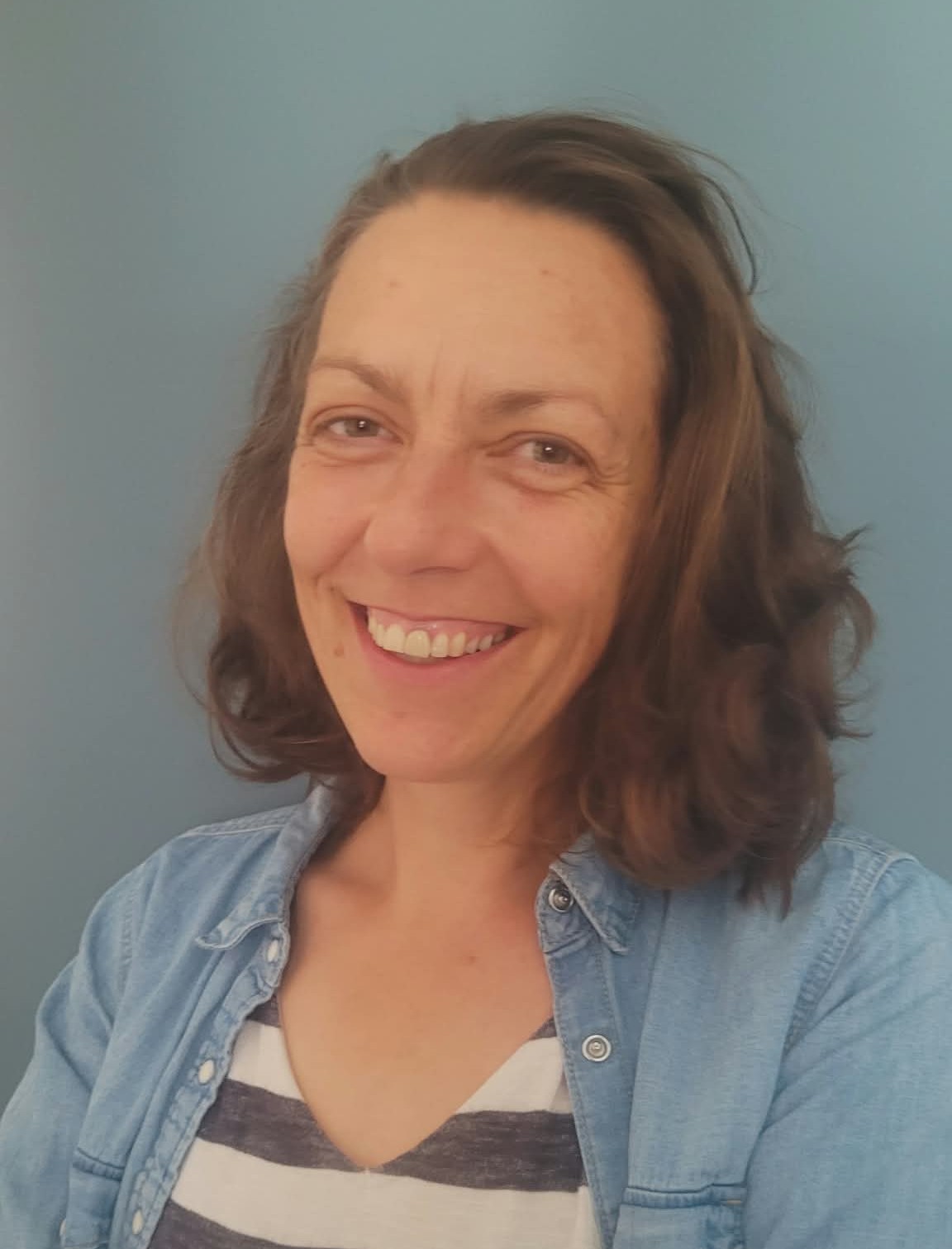 Lächelnde Frau mit kastanienbraunem, schulterlangem Haar, die eine hellblaue Jeansjacke über einem weiß-blau gestreiften Oberteil trägt, vor einem einfachen graublauen Hintergrund