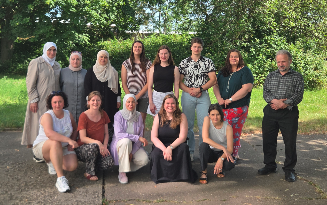 Gruppe von 13 Personen, die für ein gemeinsames Foto im Freien posieren, einige kniend, andere stehend vor grünen Bäumen