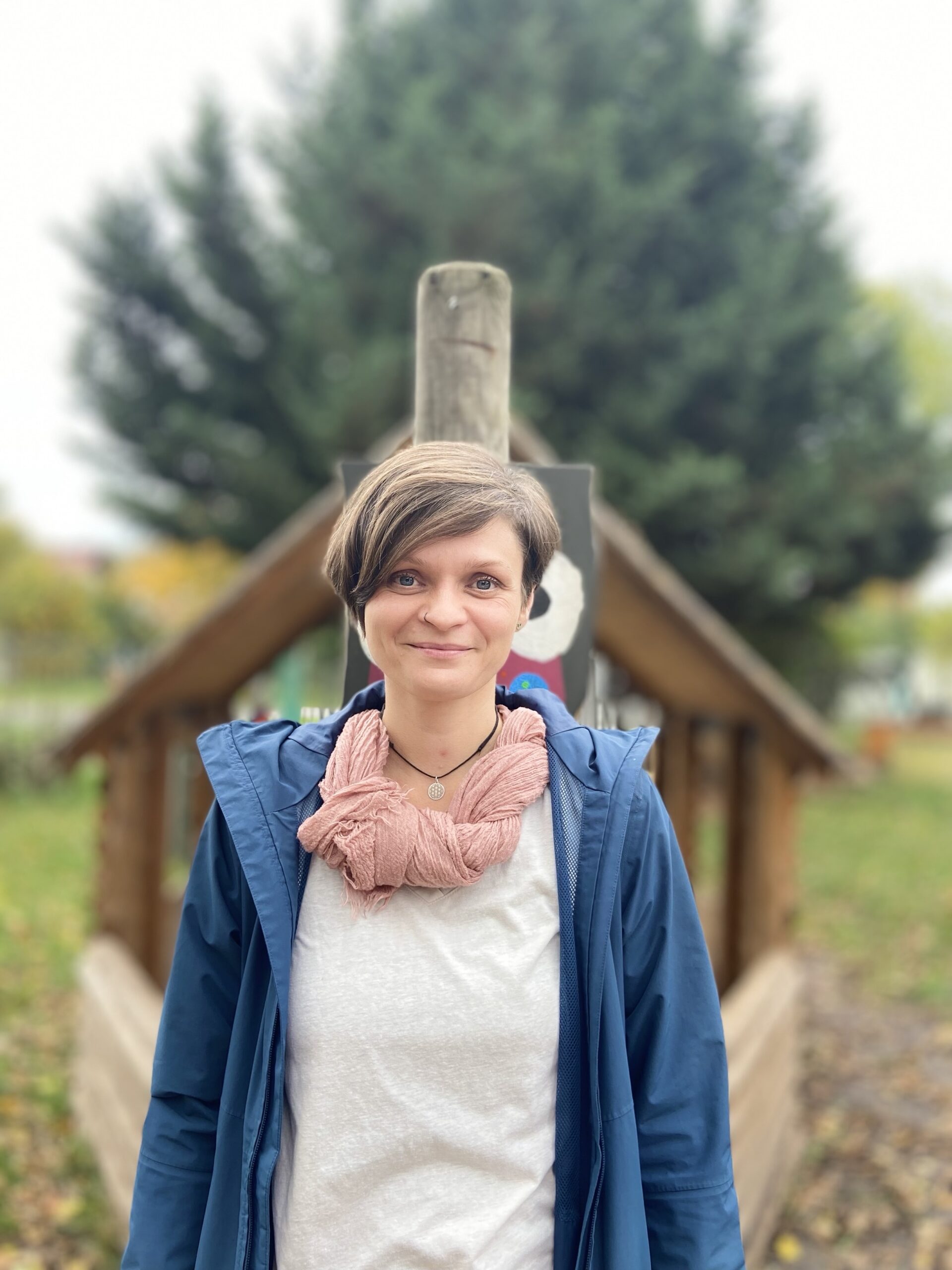 Lächelnde Person mit kurzen Haaren, blauer Jacke und rosa Schal vor Holzgerüst und Bäumen im Hintergrund