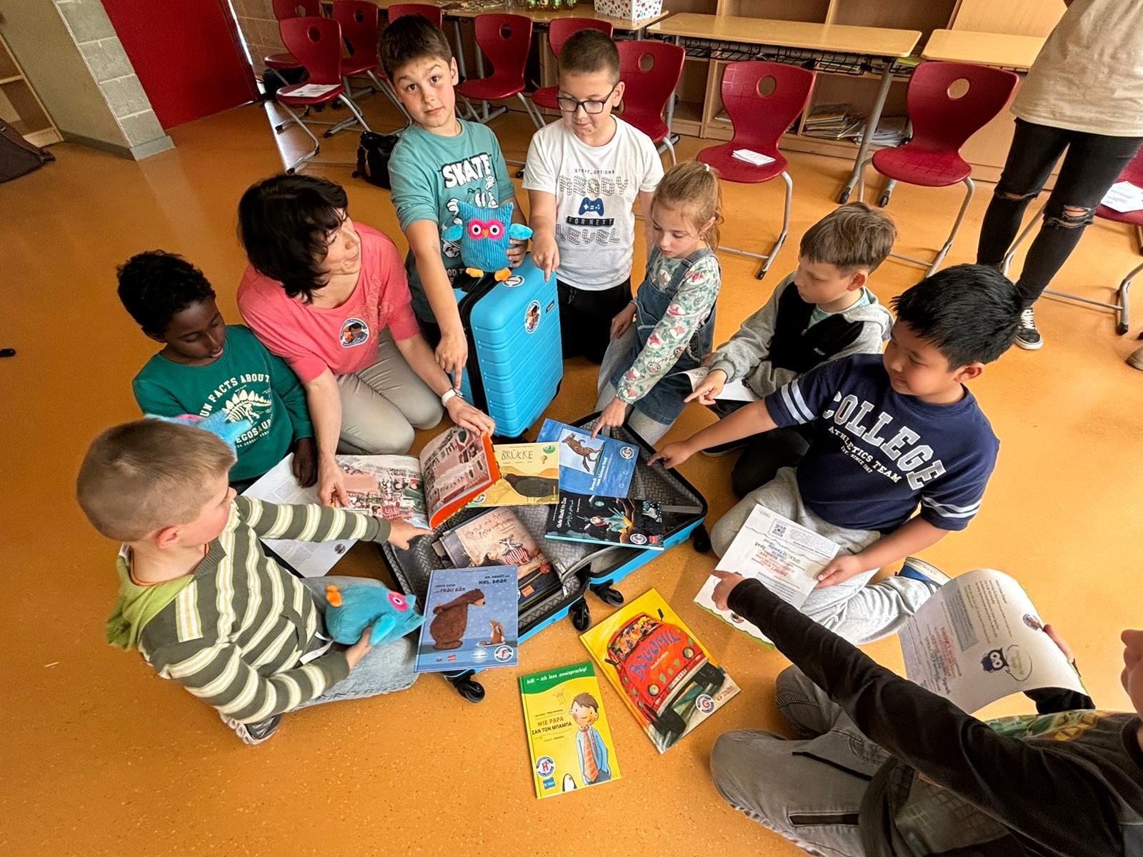 Kinder und eine Erwachsene sitzen im Klassenzimmer um einen geöffneten Koffer voller Bücher
