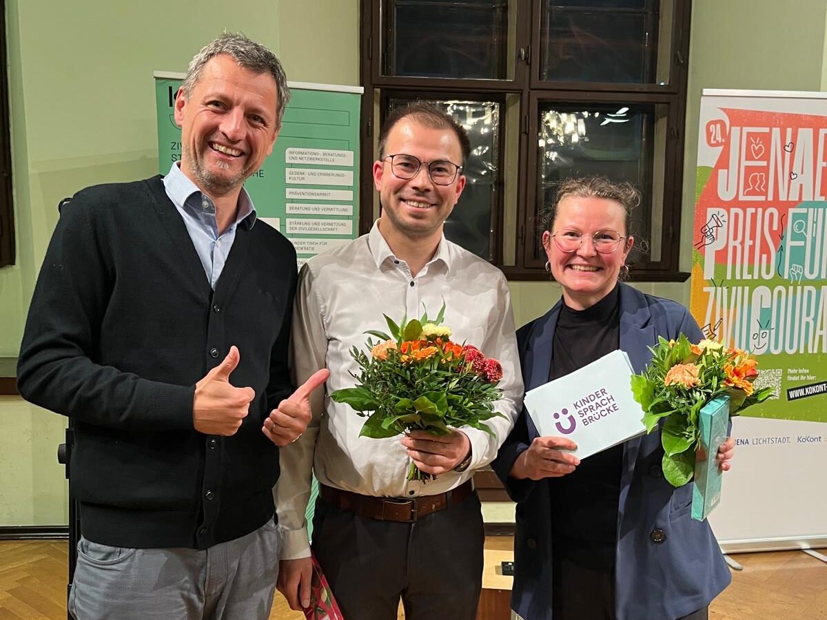 Drei lächelnde Personen, eine im Rollstuhl, mit Blumensträußen und der Urkunde „Kinder Sprach Brücke“