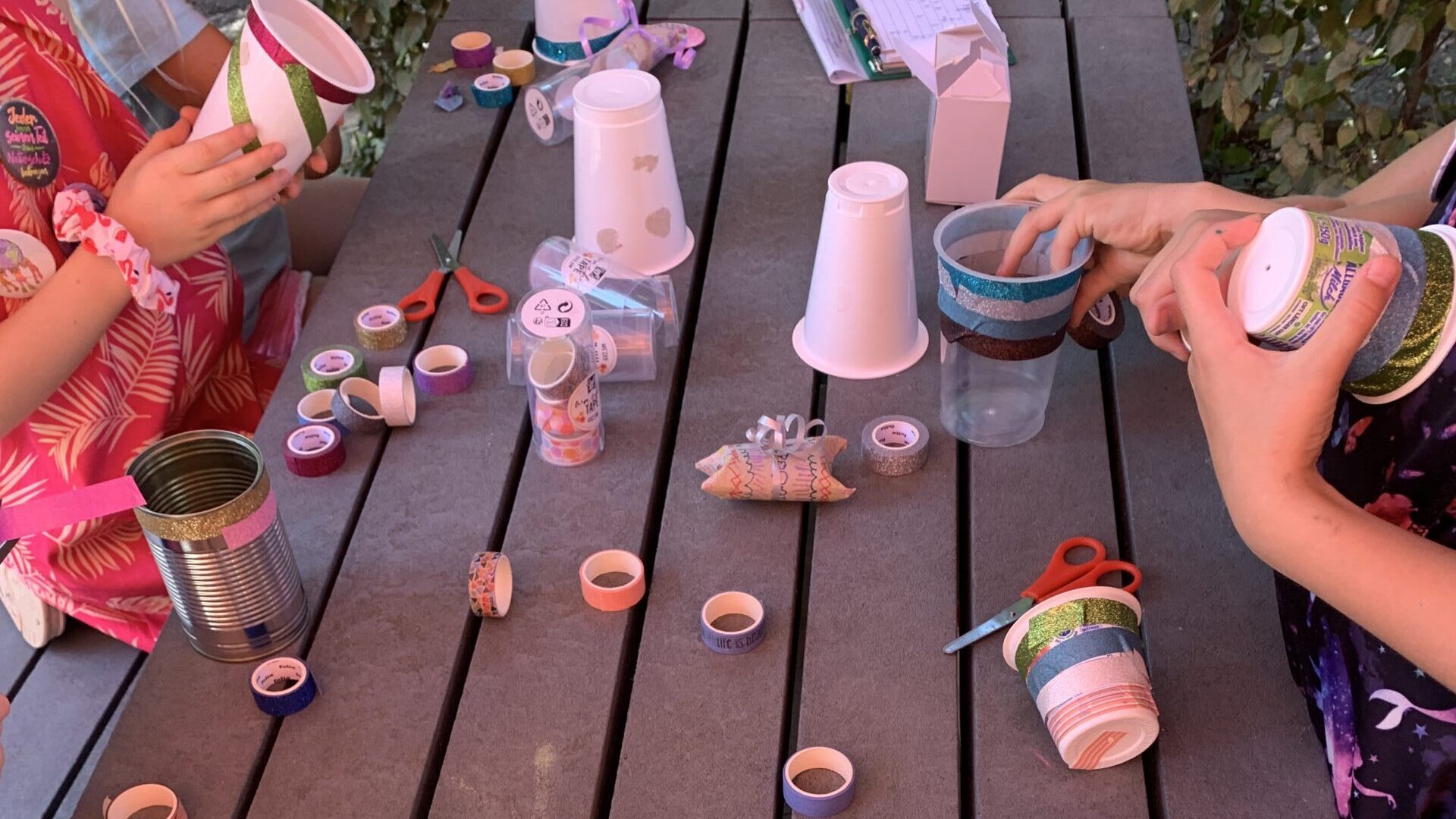 Hände von Kindern beim Basteln: Becher, Dosen und Papprollen werden mit buntem Klebeband beklebt