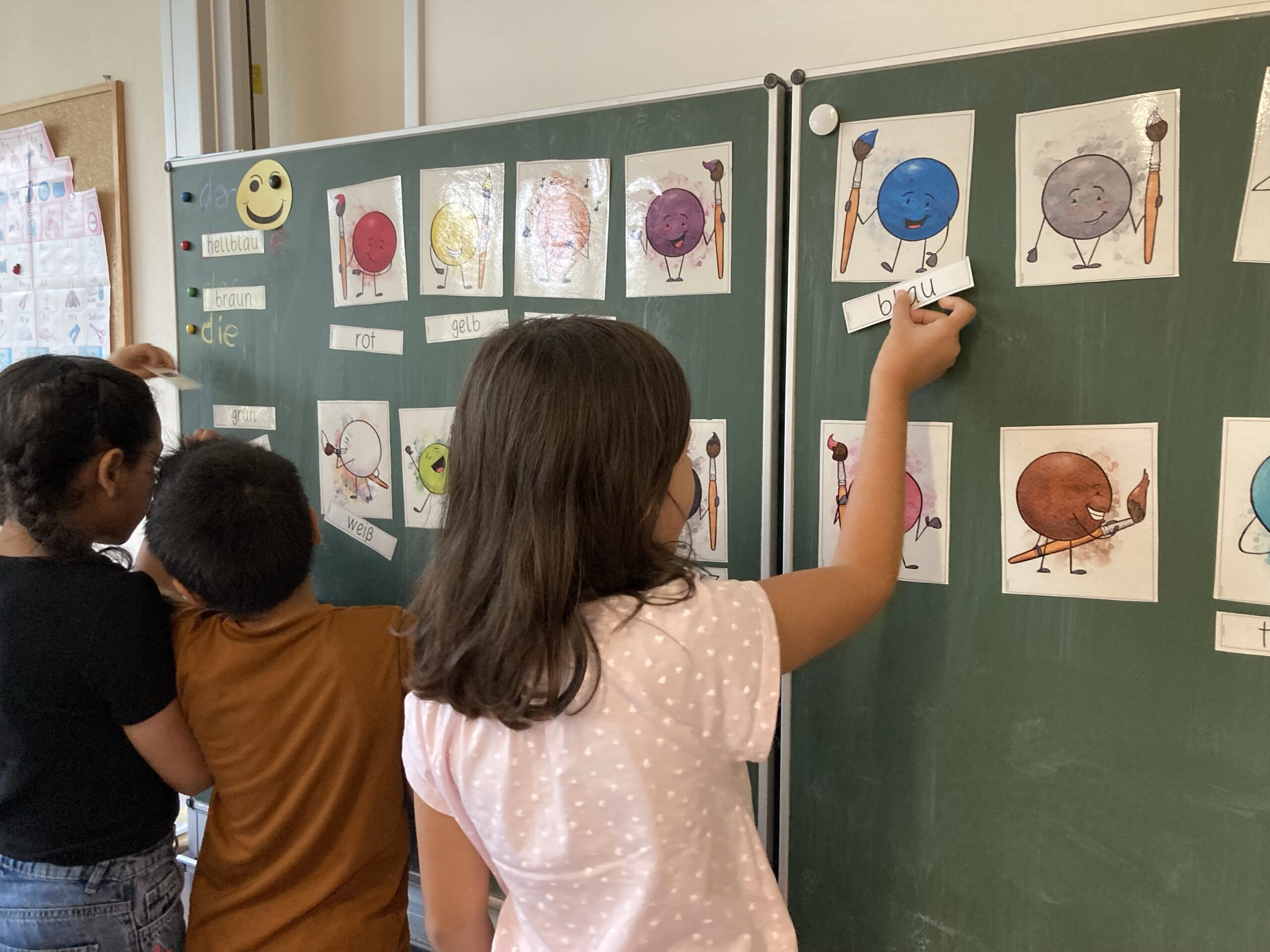 Kinder vor einer grünen Tafel mit Farbkarten; ein Mädchen hängt die Wortkarte „blau" zur blauen Figur