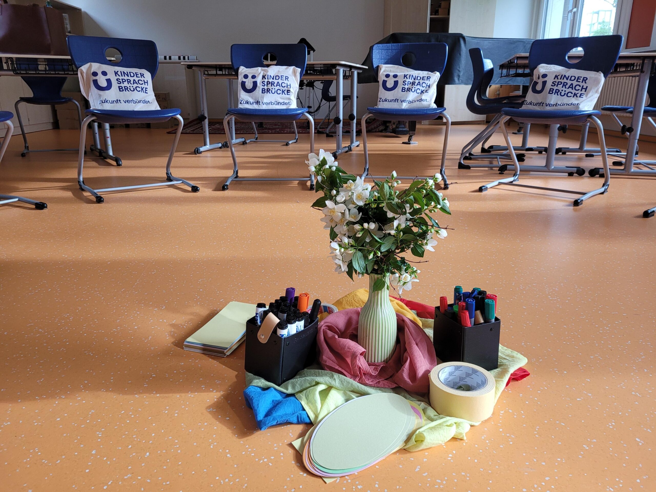Raum mit Stühlen und Taschen der Initiative Kinder Sprach Brücke, davor Blumen und Bastelmaterial auf dem Boden