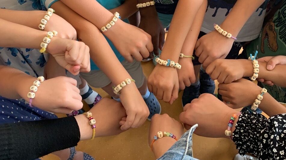 Viele Kinderhände mit bunten Buchstabenperlen-Armbändern, die sich in einem Kreis treffen