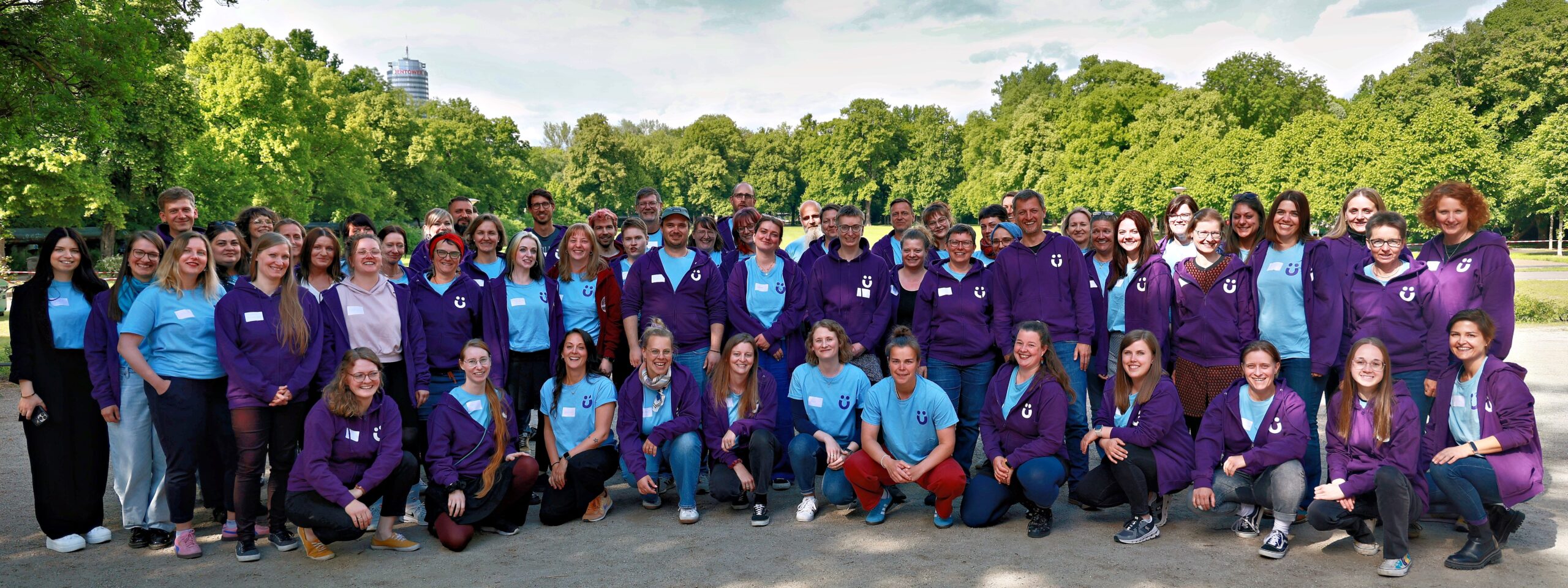 Gruppenfoto von etwa 60 Personen in lila und hellblauen Oberteilen im Freien, teils kniend und stehend