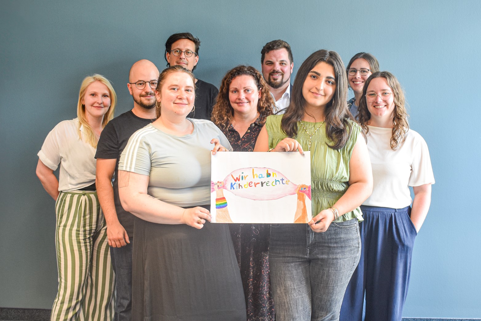 Neun Personen halten vor blauer Wand ein selbstgemaltes Schild mit der Aufschrift Wir habn Kinderrechte