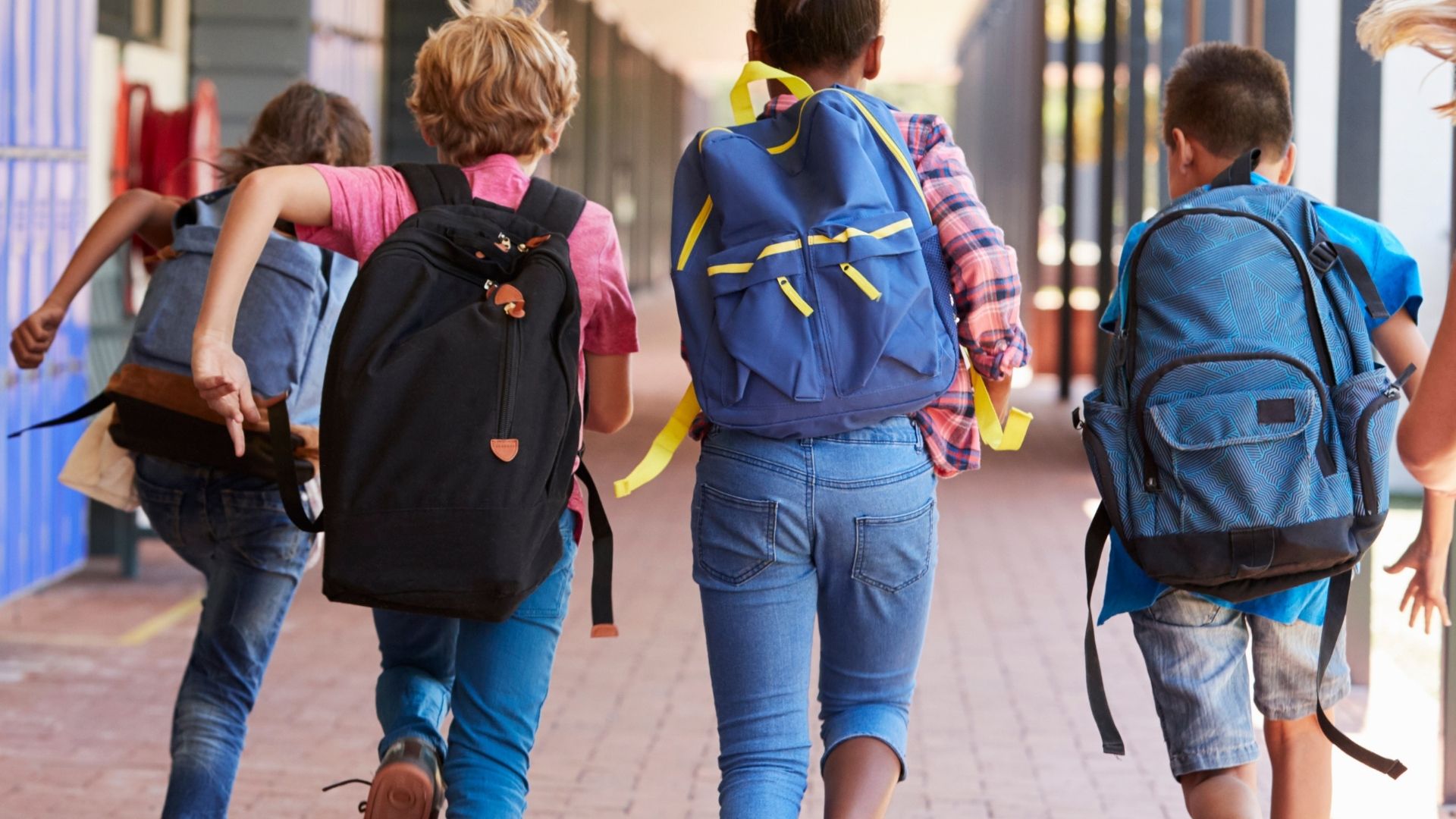 Vier Kinder mit Rucksäcken laufen von hinten gesehen über einen Gehweg