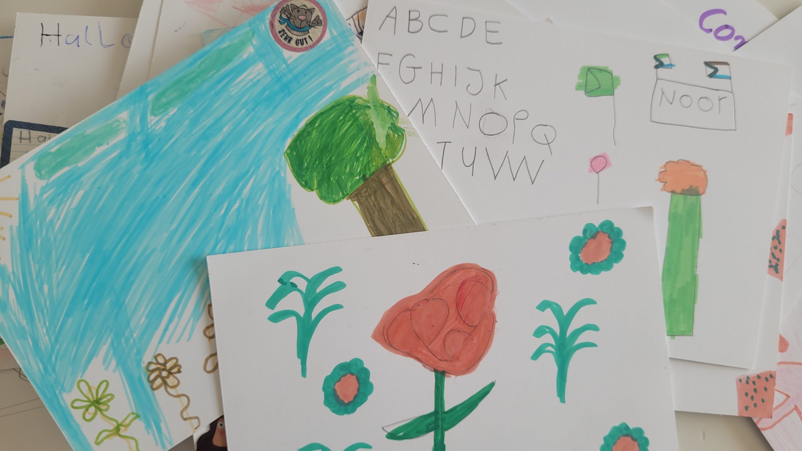 Übereinanderliegende Kinderzeichnungen mit Alphabet, Baum, Blumen und dem Namen Noor