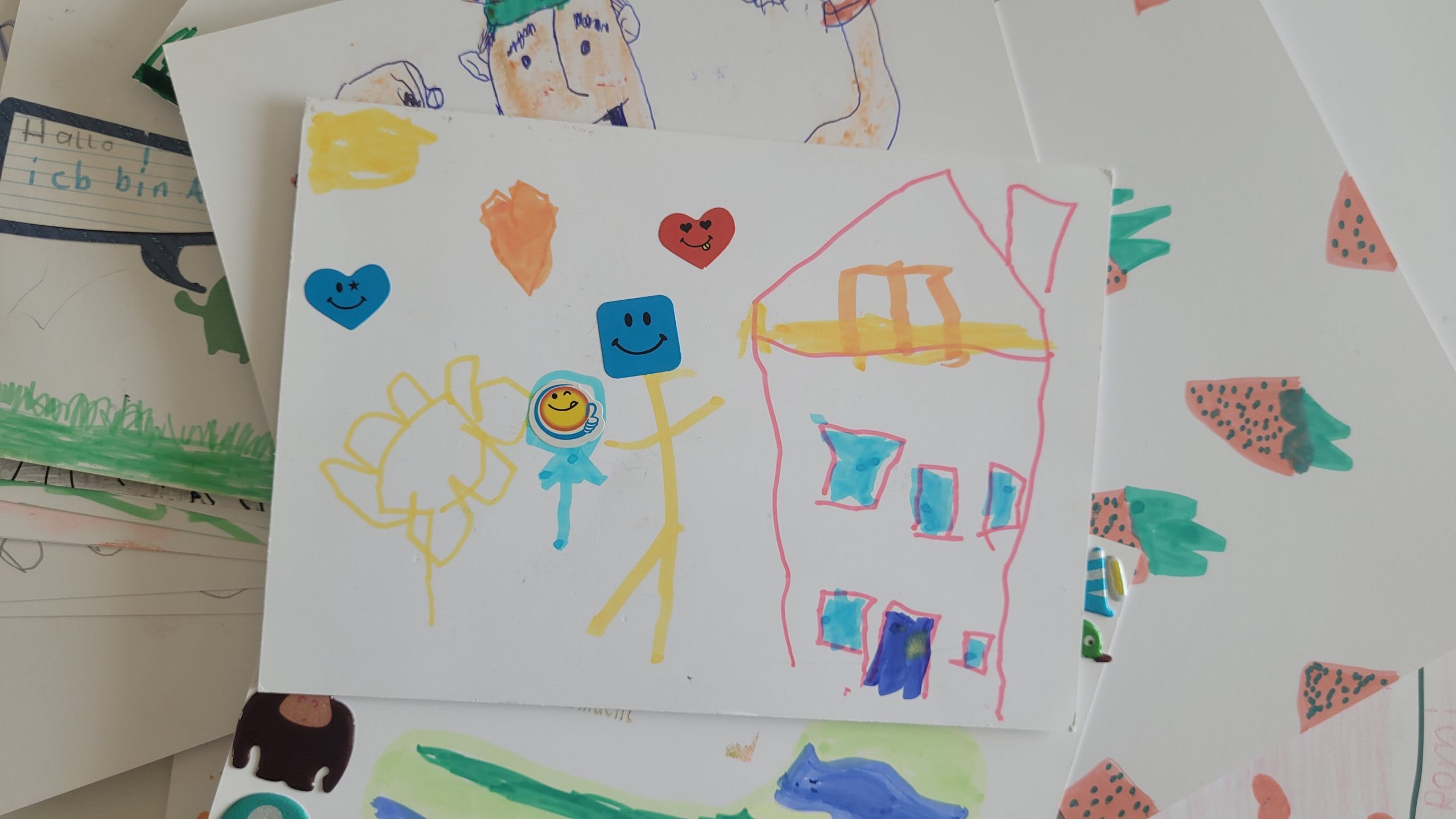 Stapel von Kinderzeichnungen, oben ein Haus, Sonne, Strichmännchen mit Stickern und Herzen