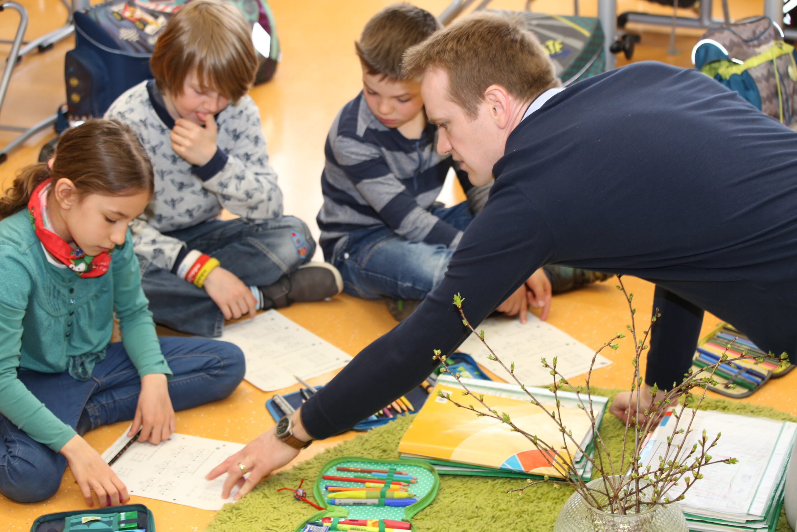 Lehrer kniet mit drei Kindern auf dem Boden und hilft ihnen bei Arbeitsblättern in einer Klasse