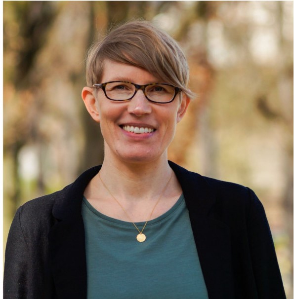 Lächelnde Frau mit kurzen blonden Haaren, Brille, schwarzem Blazer und grünem Shirt vor unscharfem Hintergrund