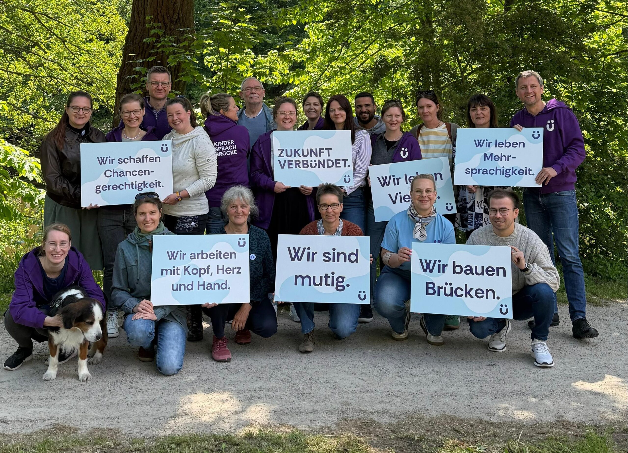 Gruppe von rund 20 lächelnden Menschen hält Schilder mit Botschaften wie Wir bauen Brücken im Grünen, vorn links sitzt ein Hund