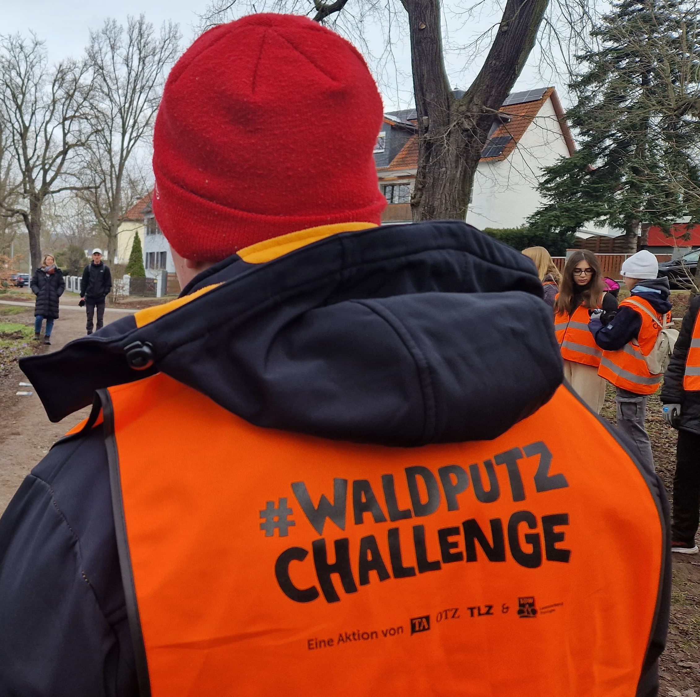 Rückansicht einer Person in orangefarbener Weste mit der Aufschrift #Waldputz Challenge, daneben weitere Helfende in Warnwesten