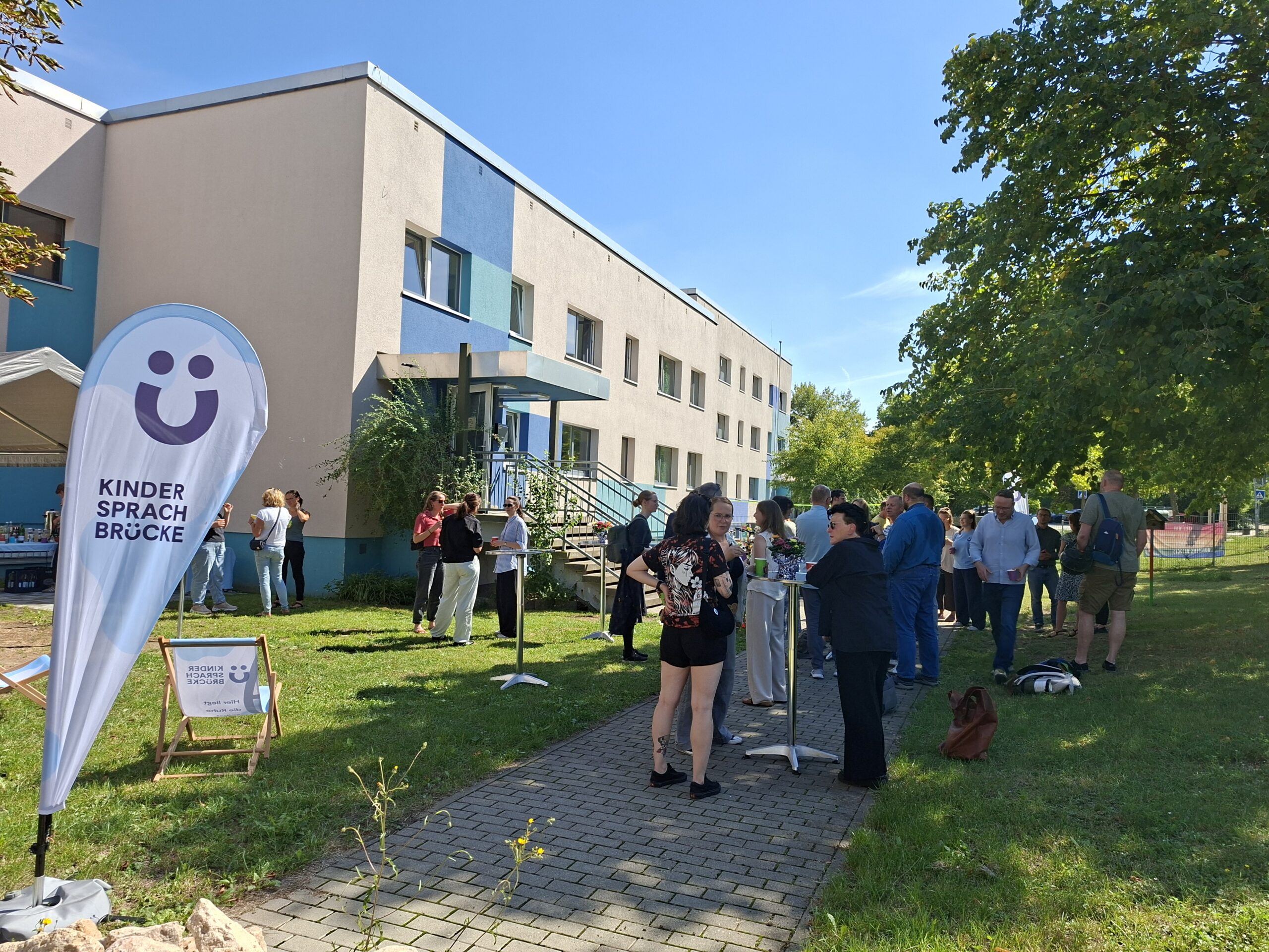 Menschen treffen sich im Garten vor einem Gebäude, daneben eine Fahne mit der Aufschrift Kinder Sprach Brücke