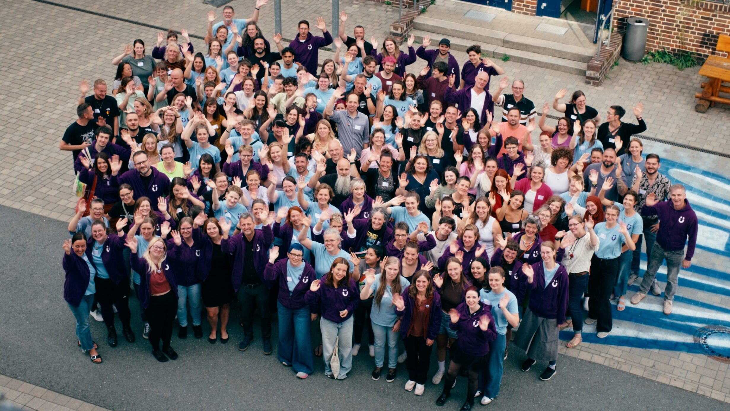 Große Gruppe von Menschen in hellblauen und violetten Shirts winkt aus der Vogelperspektive in einem Innenhof in die Kamera