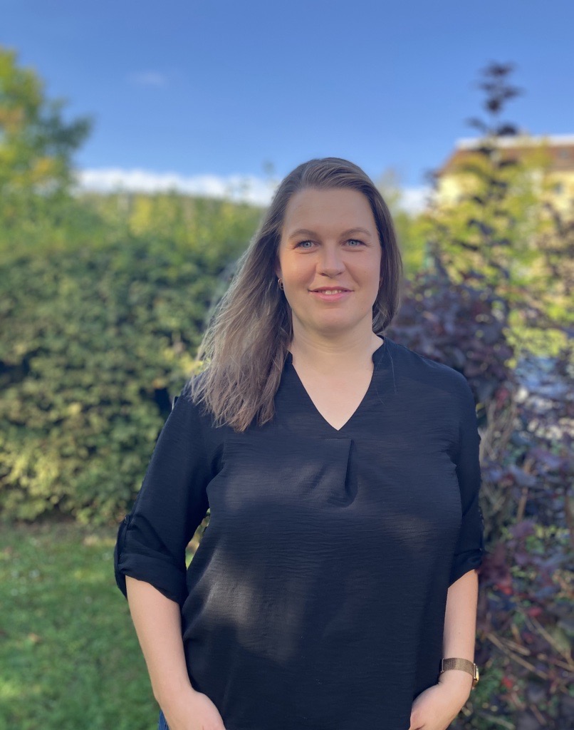 Lächelnde Frau mit braunem Haar und schwarzer Bluse mit grünen Sträuchern und HImmel im Hintergrund