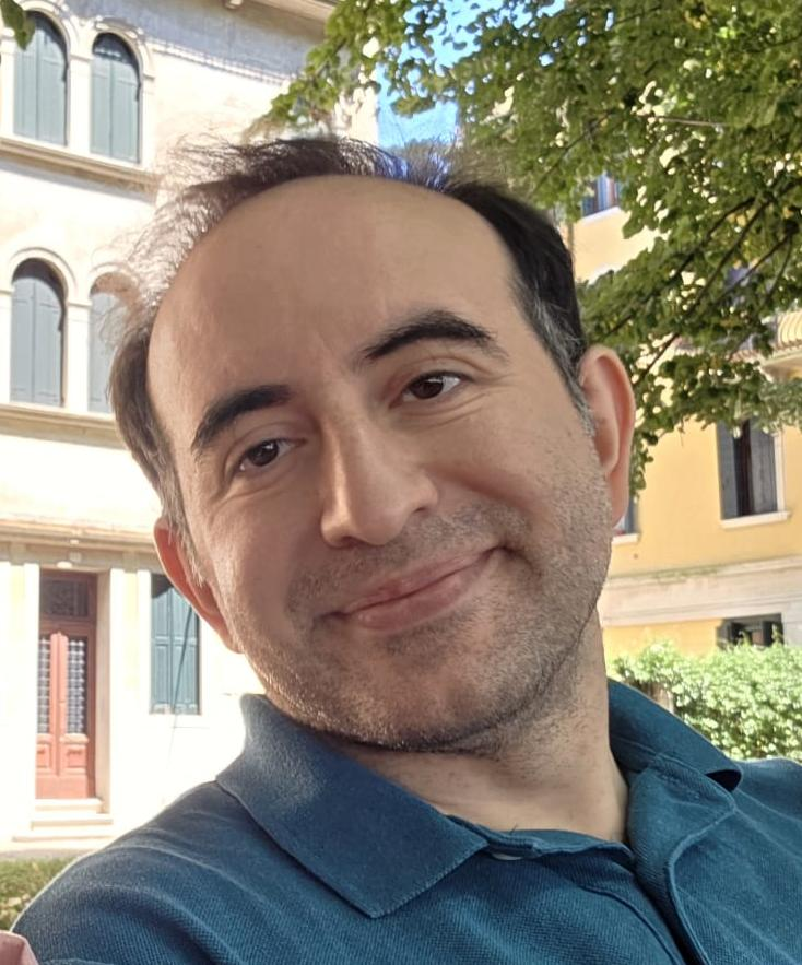 Lächelnder Mann mit schwarzem Haar und blauem Poloshirt mit grünem Baum und einem Gebäude im Hintergrund